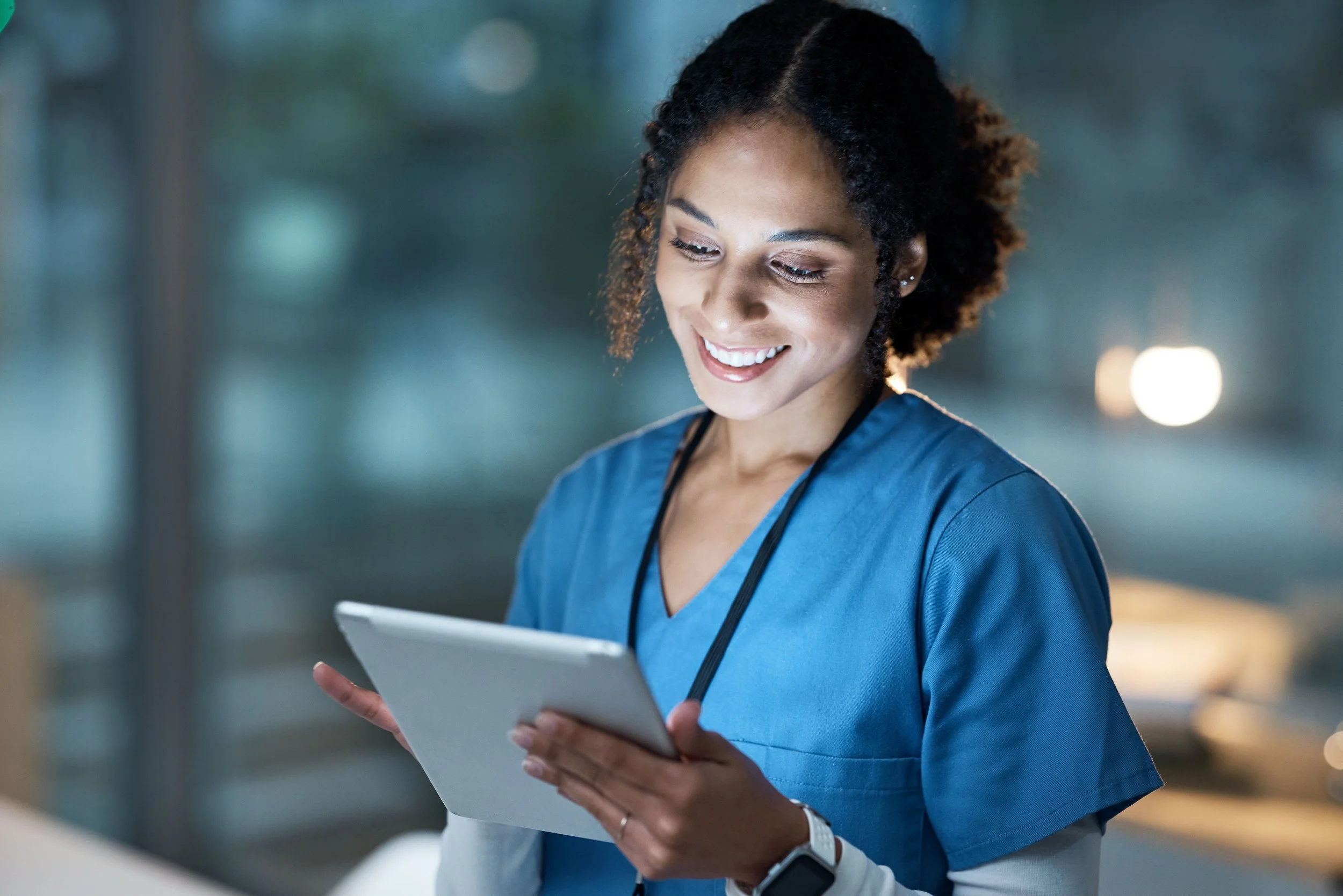 A female healthcare professional with curly hair, wearing blue scrubs and a smartwatch, is looking down and smiling while holding a tablet in her hands. The background is blurred.