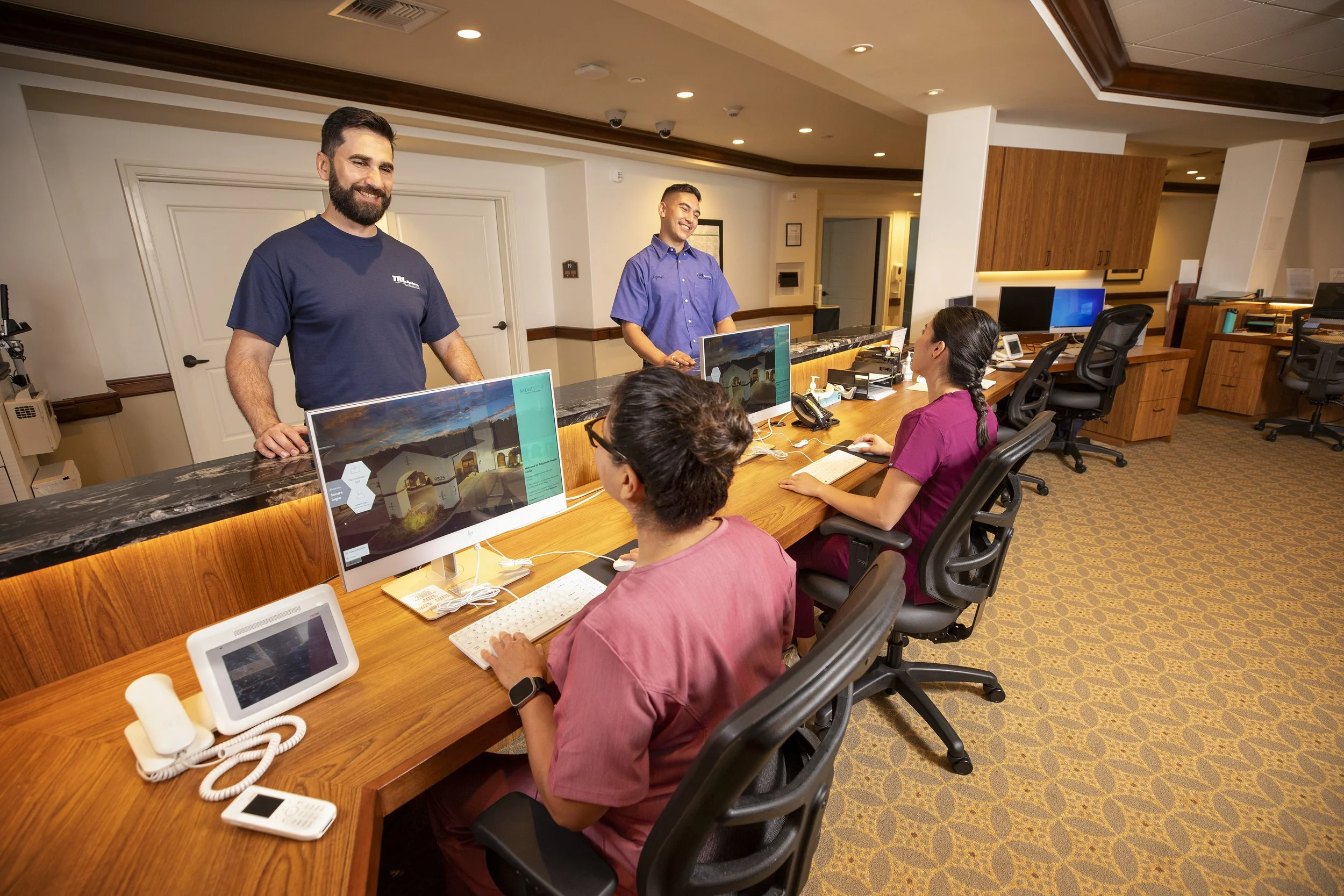 Reception desk with two smiling men standing behind it and three women seated in front, working on computers. The setting appears to be a professional office or healthcare reception area.