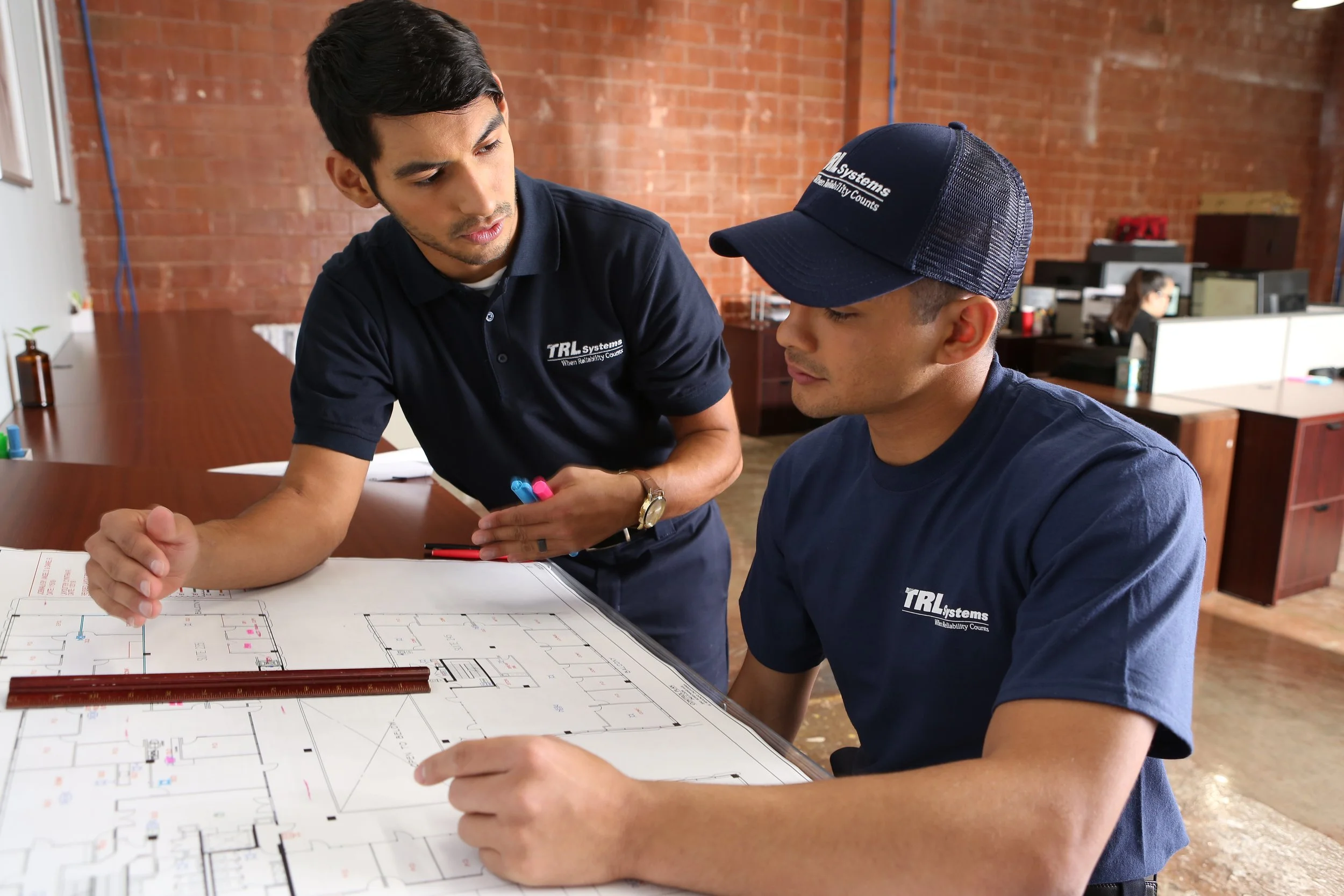 Two men working on a blueprint or floor plan in an office with exposed brick walls. One man is standing and pointing at the plan, while the other is seated, wearing a cap. Both men are wearing dark blue shirts with a company logo.