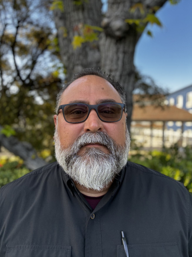 A man with sunglasses and a beard standing outdoors in front of a tree and a building.
