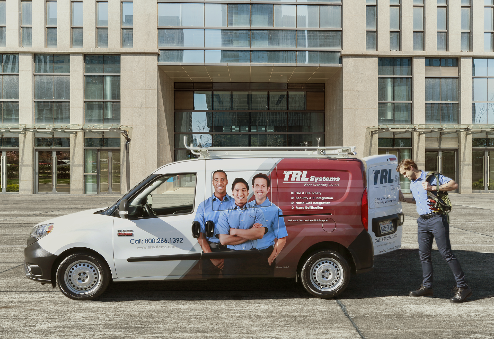 A company vehicle parked outside a modern office building with large glass windows. The van features images of three smiling employees and information about their services, including safety, security, and maintenance, with a contact phone number. A person dressed in business casual attire is walking past the van, carrying a backpack and a helmet.