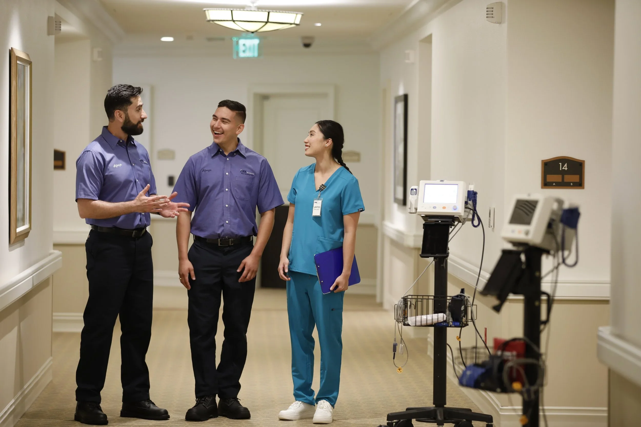 Three healthcare professionals in a hospital corridor engaging in a conversation. Two men wearing purple shirts with name tags and a woman in teal scrubs holding a clipboard are smiling and talking.