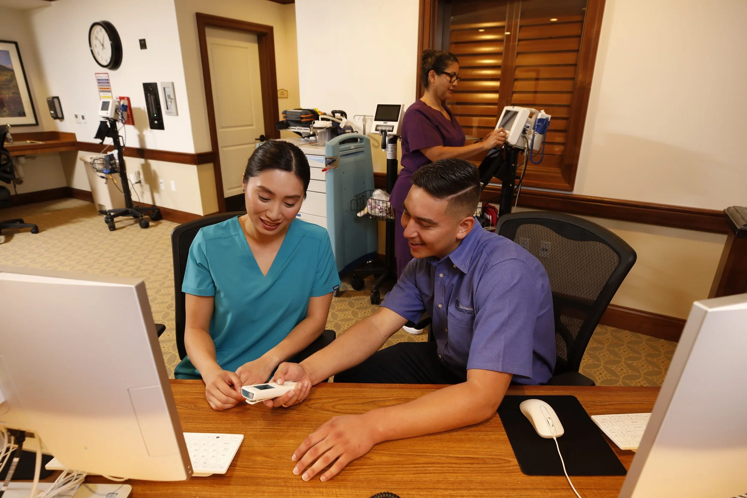A healthcare professional and a patient sitting at a desk in a medical office, looking at a blood pressure monitor. A medical staff member is in the background preparing equipment.