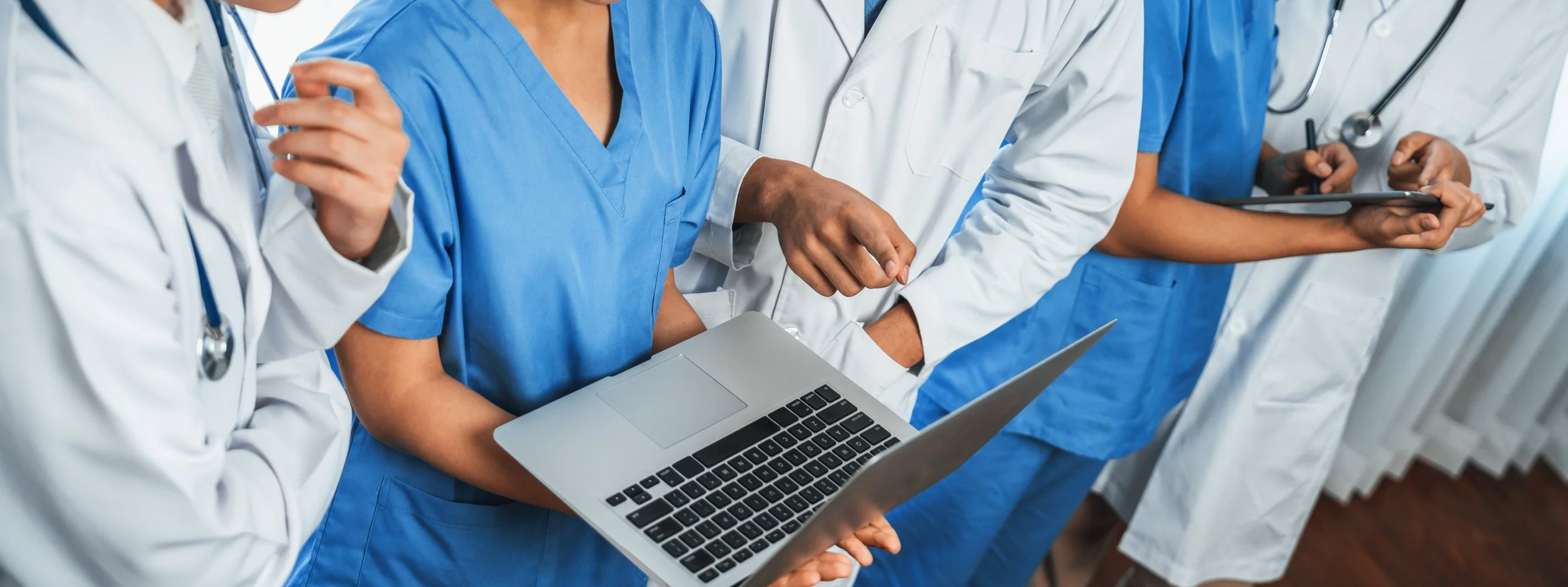 Group of healthcare professionals in scrubs and lab coats gathered around a laptop and tablet.