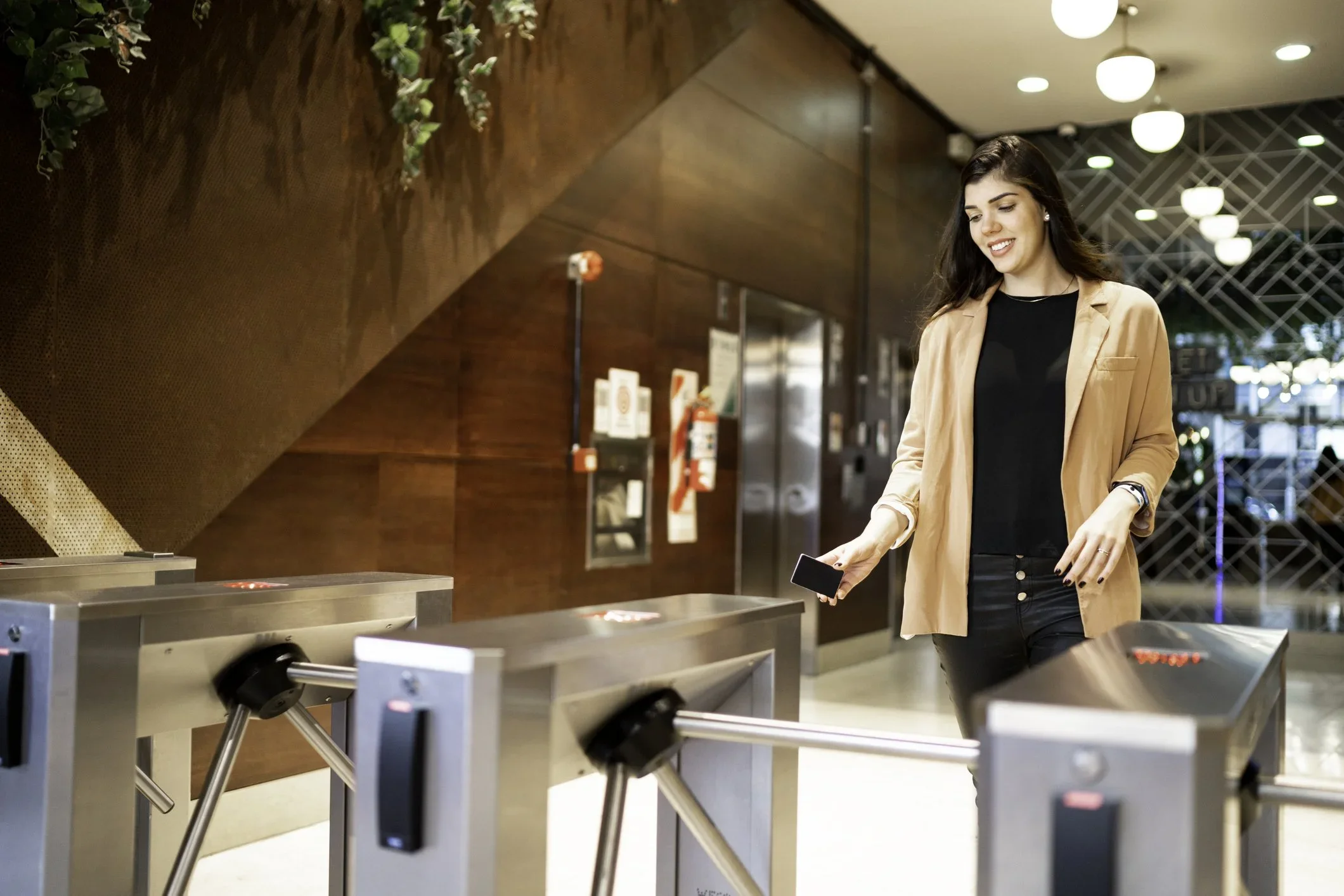 A young woman with dark hair, wearing a light brown blazer and black top, is using her phone near a row of metal turnstiles at a modern indoor location.