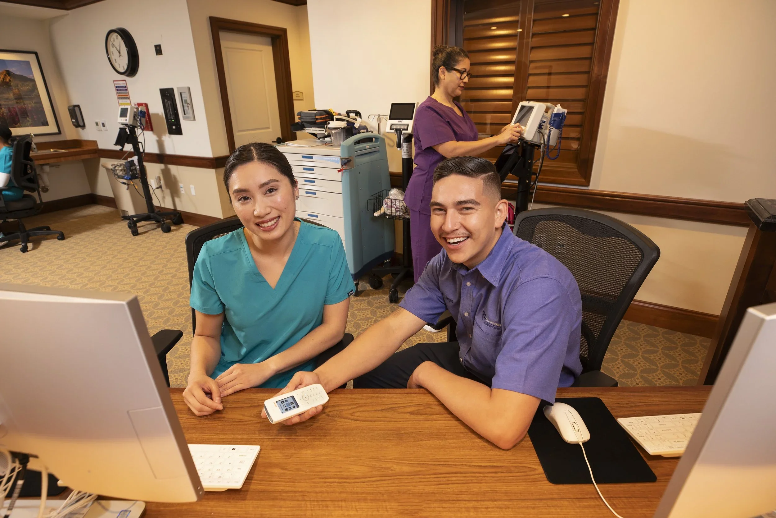 Two healthcare workers sit at a desk with computers in a medical office, smiling at the camera. A nurse in teal scrubs and a man in a purple shirt are in the foreground, while a nurse in purple scrubs works on a monitor in the background. Medical equipment and supplies are visible around the room.