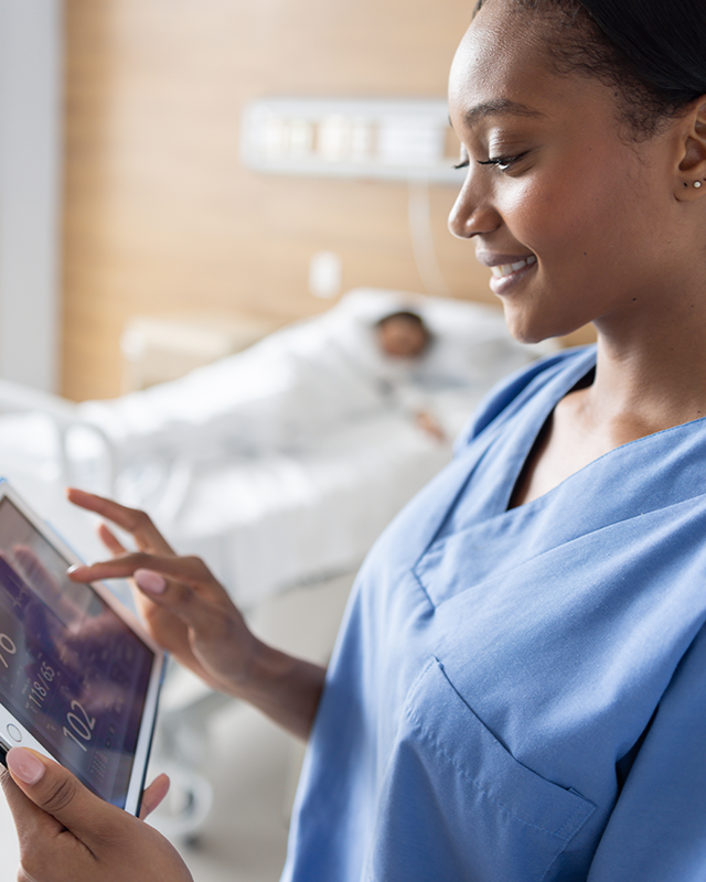 Nurse in blue scrubs smiling while looking at a tablet in a hospital room with a patient in bed in the background.