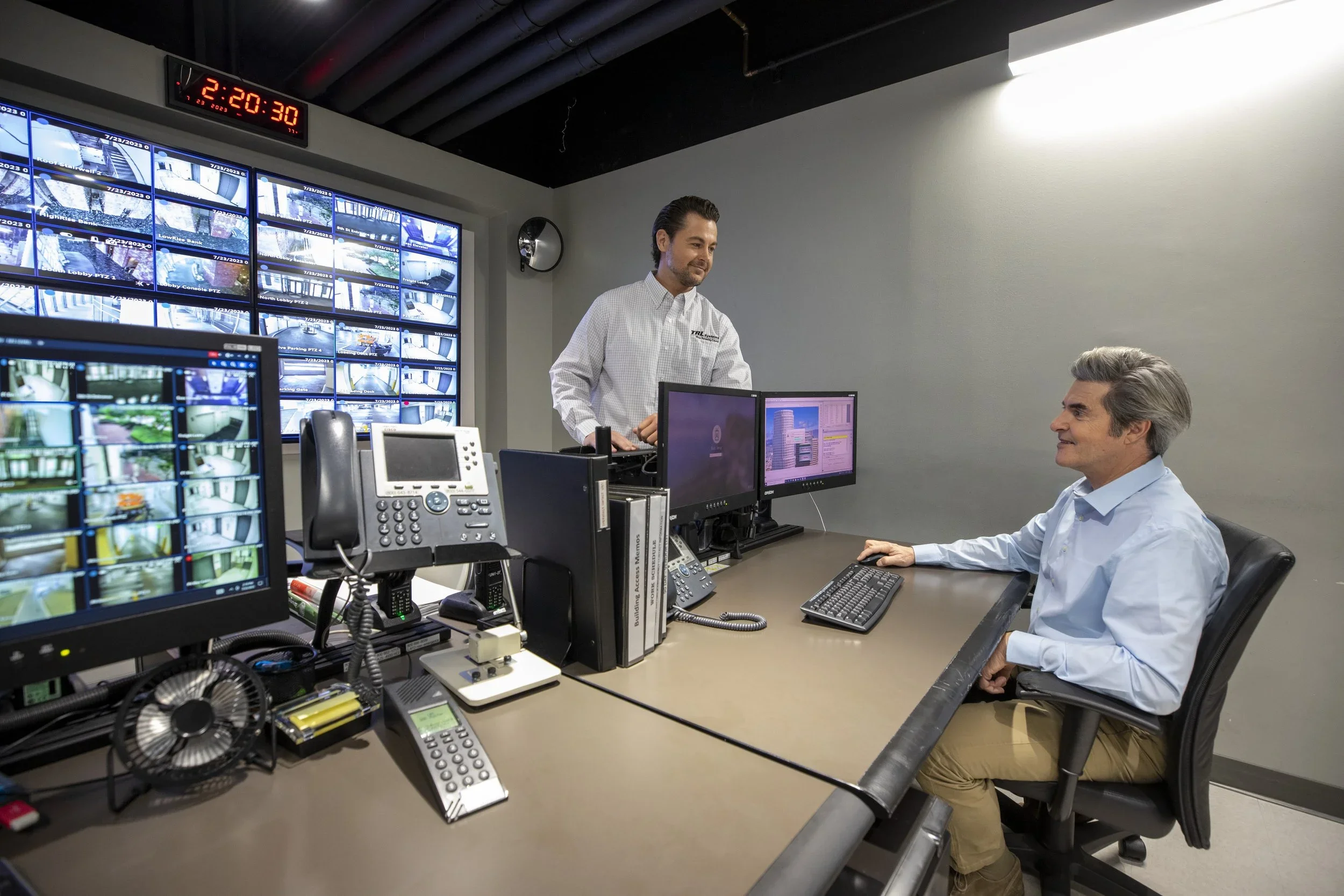 A control room with multiple computer monitors displaying security camera feeds. Two men are working; one is sitting at a desk using a computer, while the other is standing and engaging with him. The room has a modern, professional atmosphere.