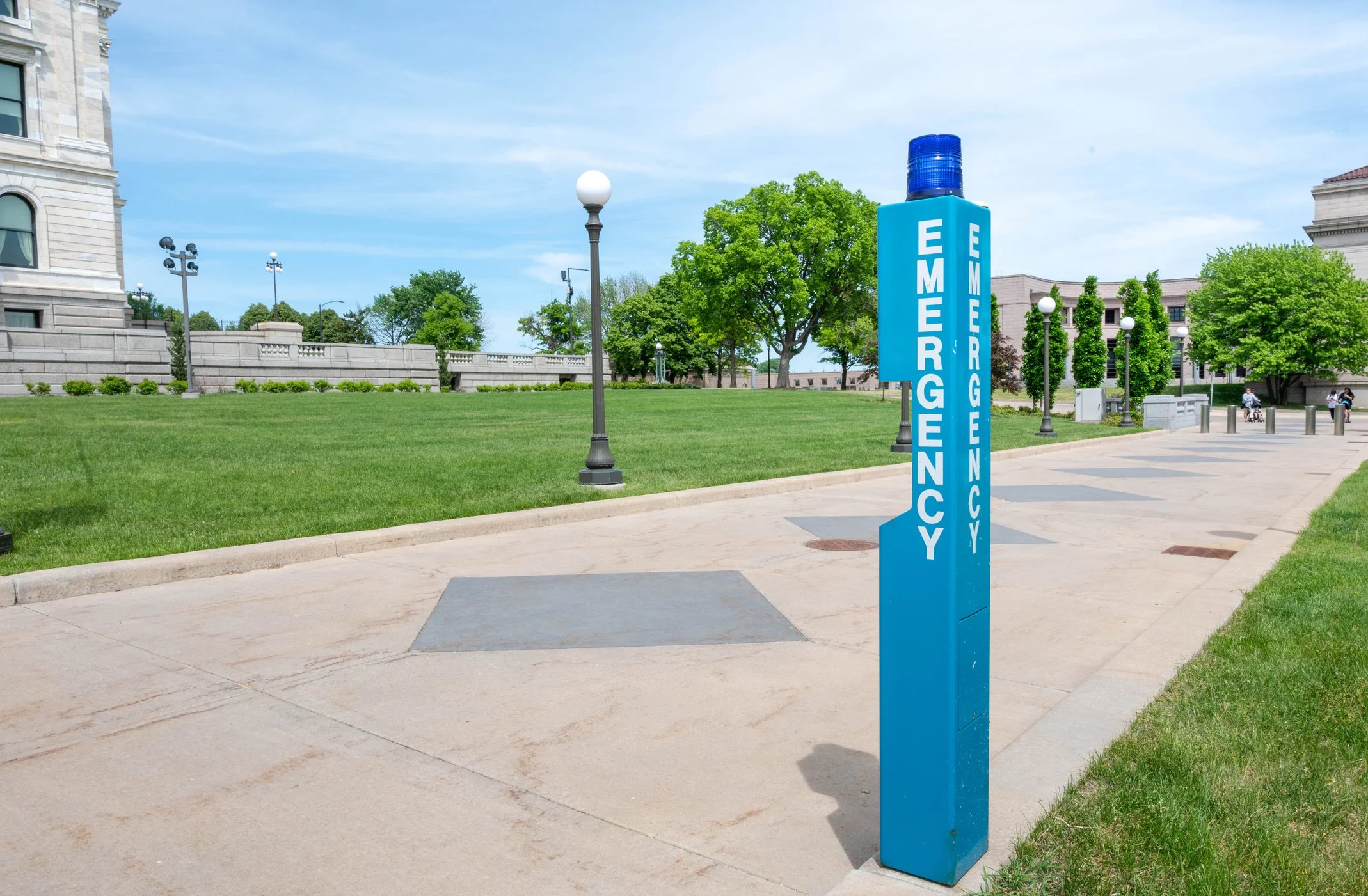 Blue emergency call button with a blue light on top, located on a sidewalk near a park and government building, with trees and benches in the background.