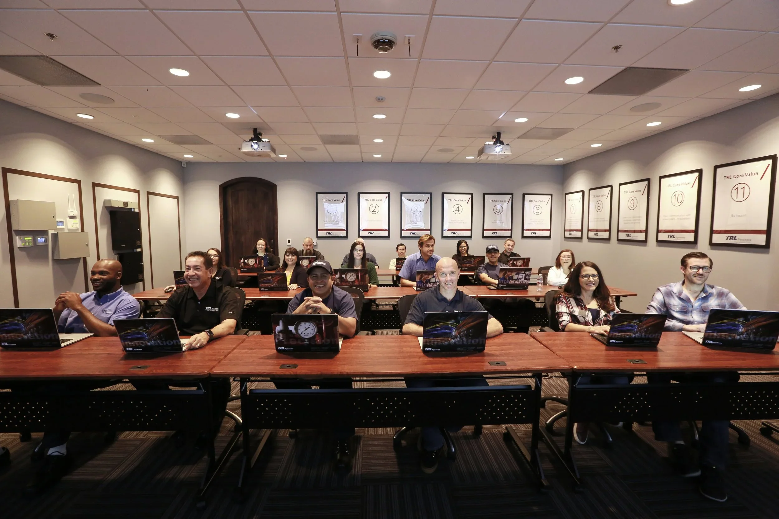 A diverse group of people sitting in a classroom or conference room, smiling and looking at the camera, with laptops open on the table in front of them. There are posters on the wall with the title ‘TRL Core Value’ and numbered points, and the room has a ceiling with lights and two projectors mounted on it.