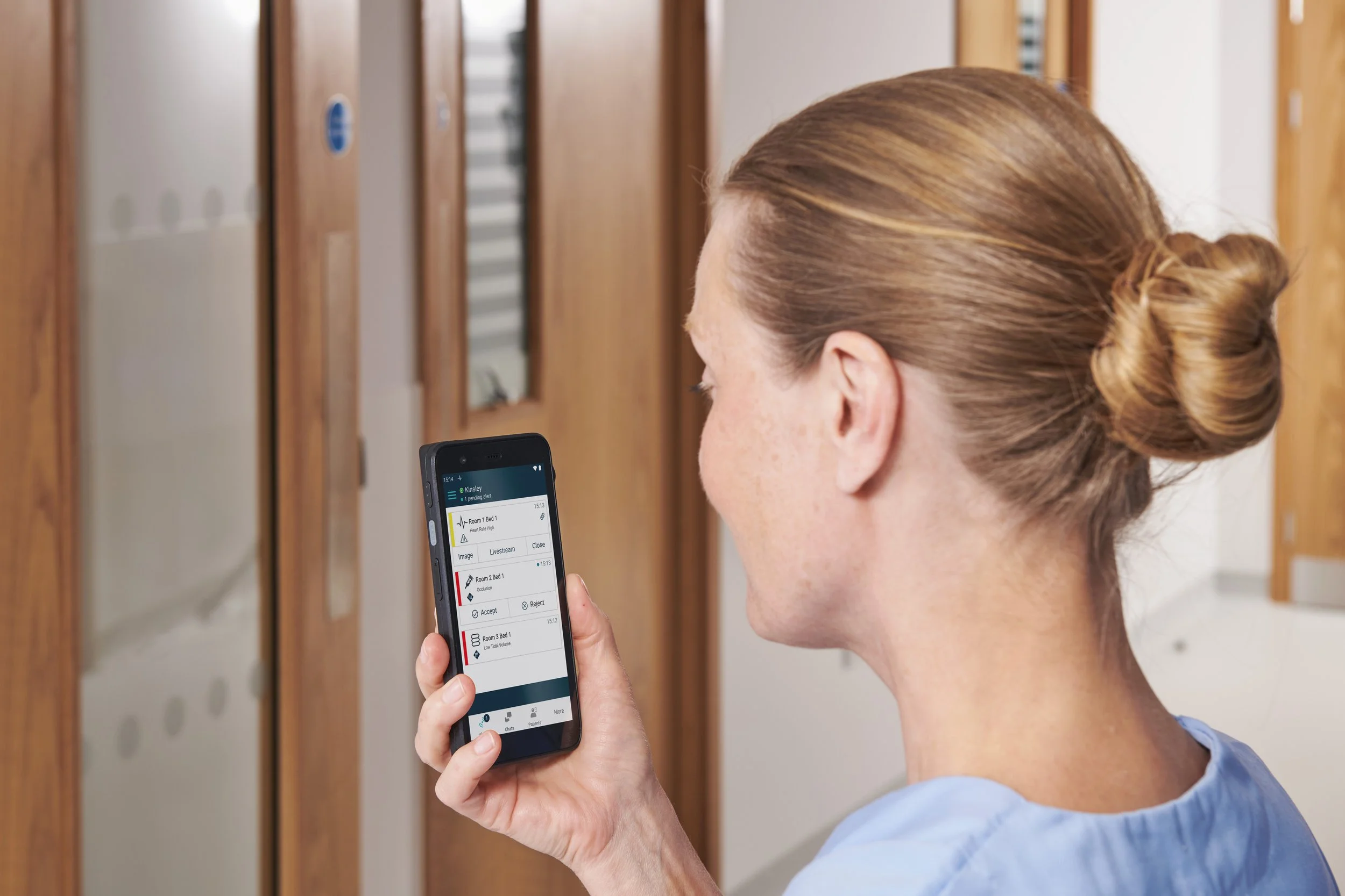 A woman with brown hair tied in a bun looking at her phone in front of bank lockers.