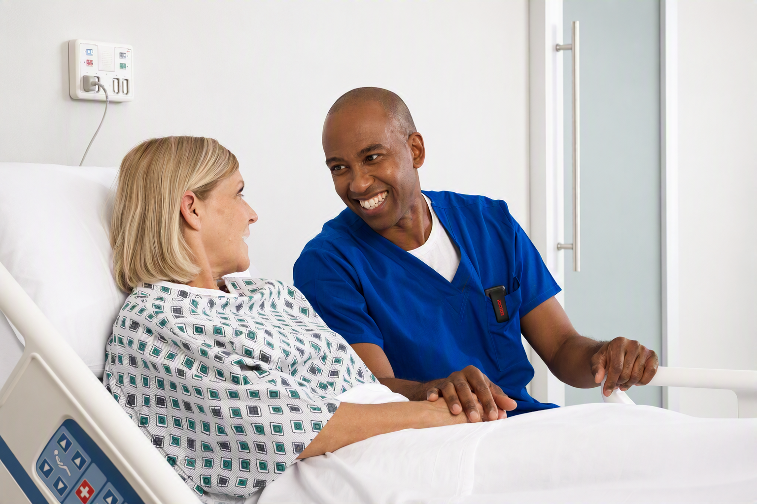 A healthcare professional smiling and talking to an older woman in a hospital bed, holding her hand. The woman is in a hospital gown, and the setting is a hospital room.