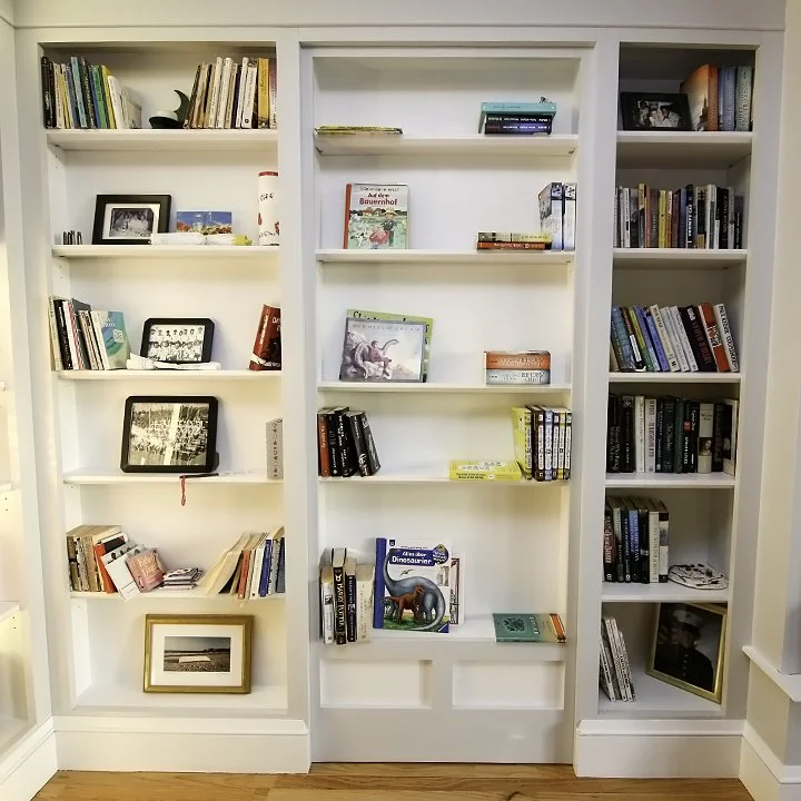 Three white bookshelves filled with various books, framed photos, and decorative items in a room with a wooden floor.