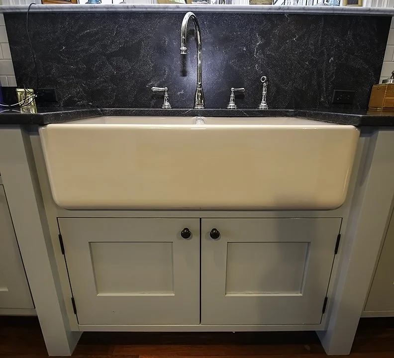 A farmhouse-style kitchen sink with a cream-colored apron front and a black granite countertop, set above a white cabinet with black knobs.