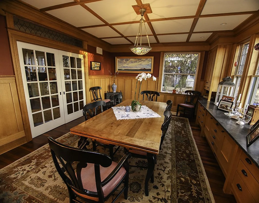 A dining room with wooden paneling, a rectangular wooden table surrounded by six black chairs, a decorative rug, and a sideboard. The room features large windows, a sliding glass door, and a ceiling light fixture with a decorative glass shade.