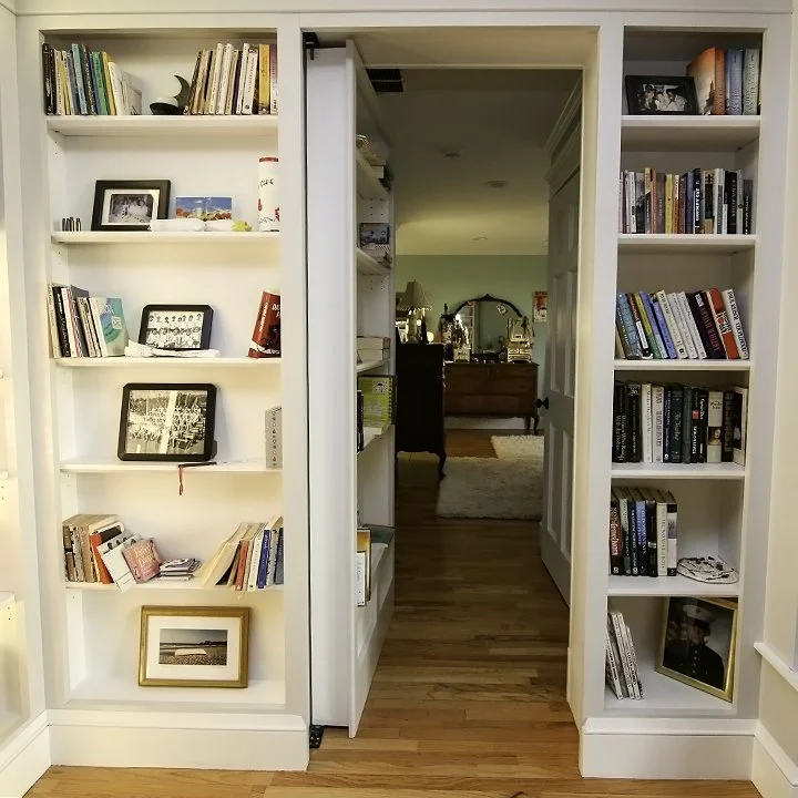 A doorway with white built-in bookshelves on each side, filled with books and framed photos, leading into a room with a mirror, dresser, and living area with wooden flooring.