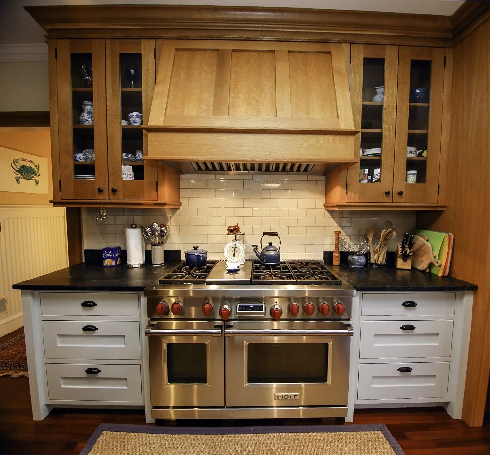 Kitchen with a stove, wooden cabinets with glass doors, black countertops, and a white subway tile backsplash. Items on the counters include a scale, kettle, spice bottle, cutting boards, and utensils. Wooden range hood above the stove.