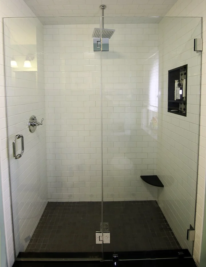 A modern walk-in shower with white subway tile walls, a rainfall showerhead, and a built-in black shelf for toiletries, enclosed by glass.