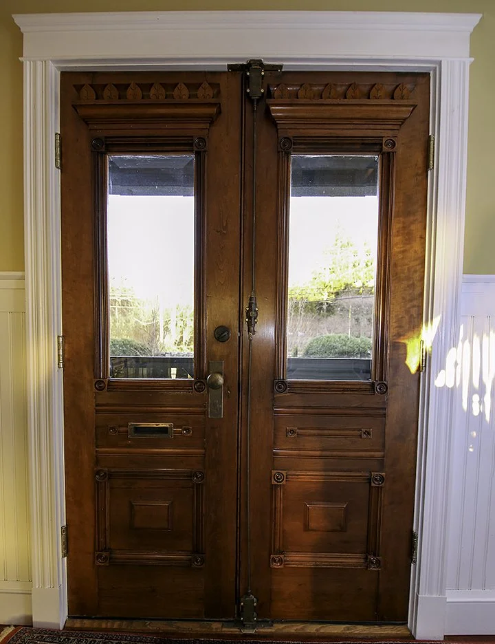A pair of wooden front doors with decorative carved panels and glass panes, surrounded by white trim, with sunlight shining on the right side.