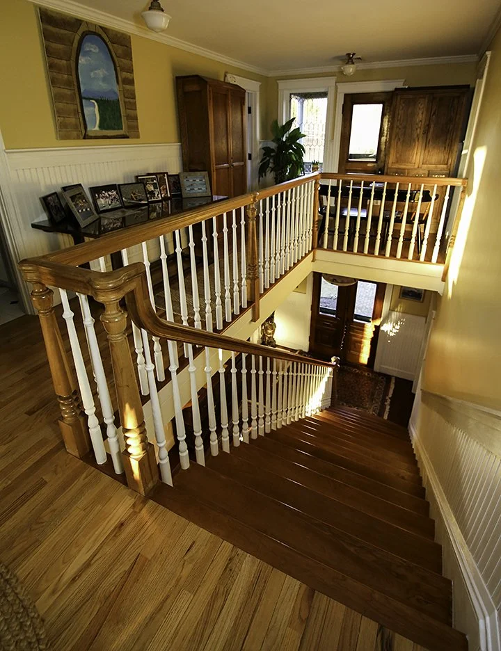 Interior view of a two-story house staircase with wooden steps and custom white and wood handrails, overlooking the entryway and front door, with yellow walls, a small table with family photos, a wooden cabinet, and a large potted plant near a window