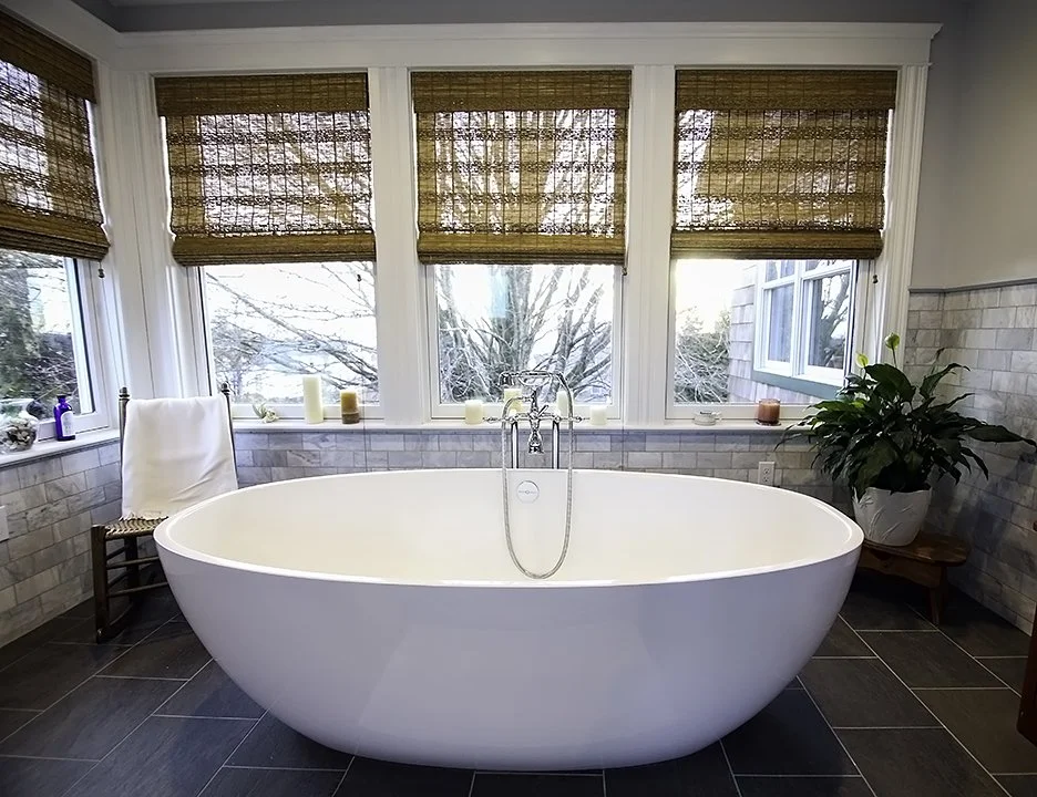 A white freestanding bathtub in a bright bathroom, surrounded by large windows with bamboo blinds, a potted plant on a small wooden table, and candles on the ledge.