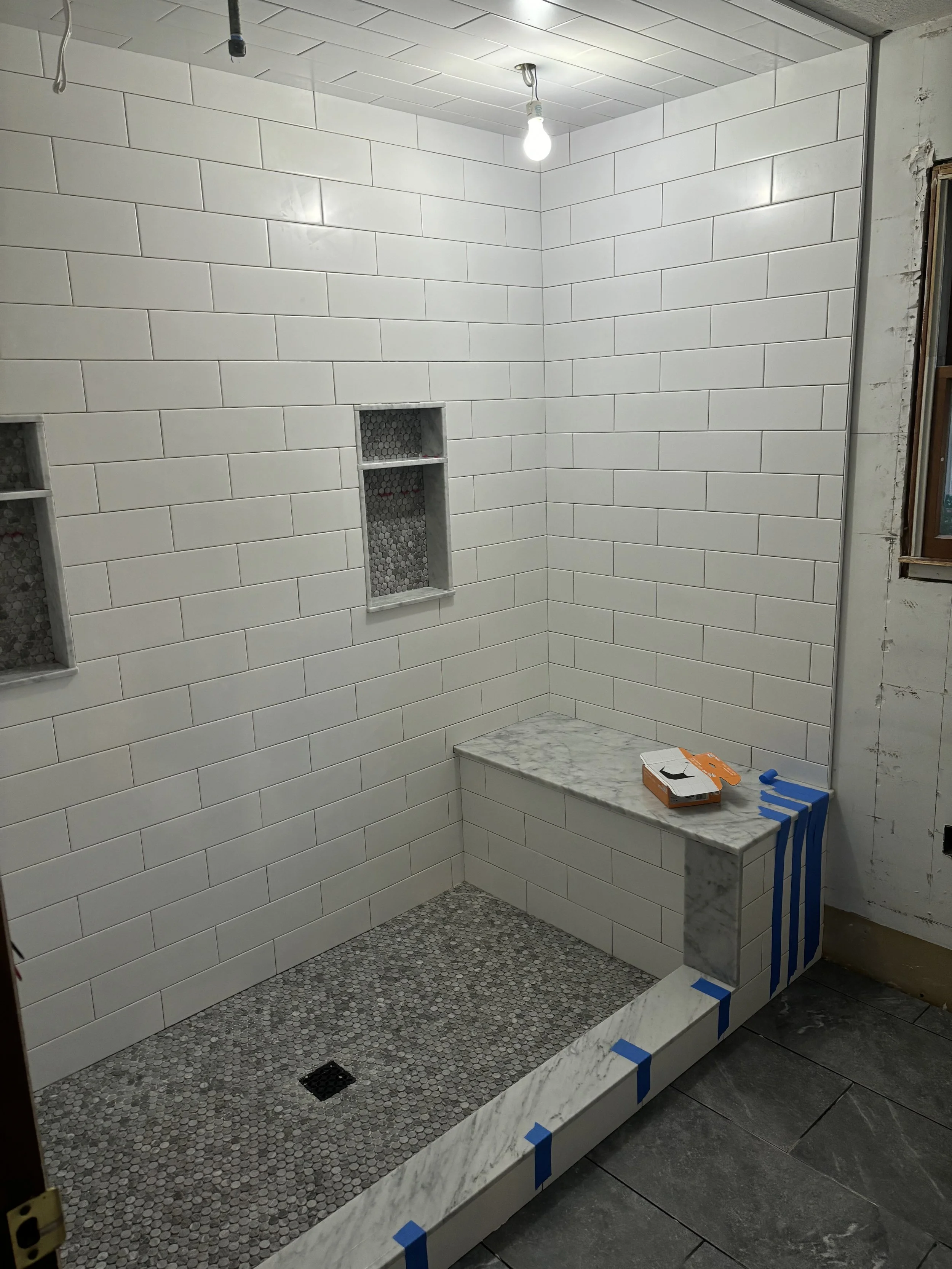 A partially finished walk-in shower with white subway tile walls, gray mosaic tile in the shower niche and floor, a marble bench, and construction materials including blue painter's tape and a box, with a single lightbulb hanging from the ceiling.