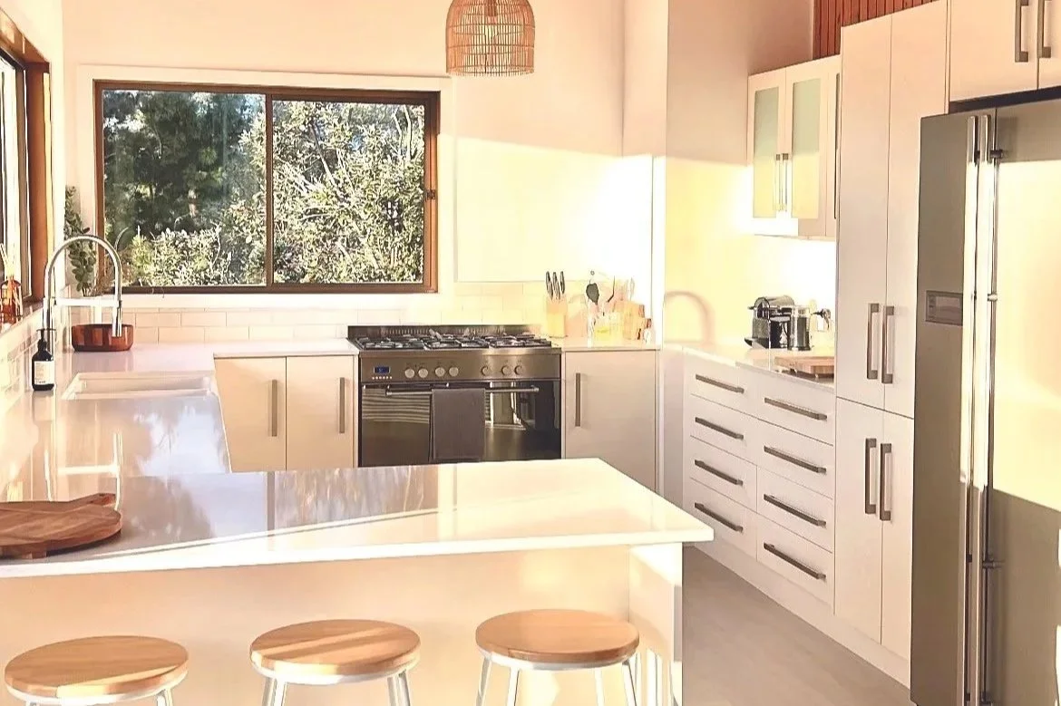 Bright kitchen with white cabinets, a large window, a stainless steel oven, a white kitchen island with three wooden stools, and a refrigerator.