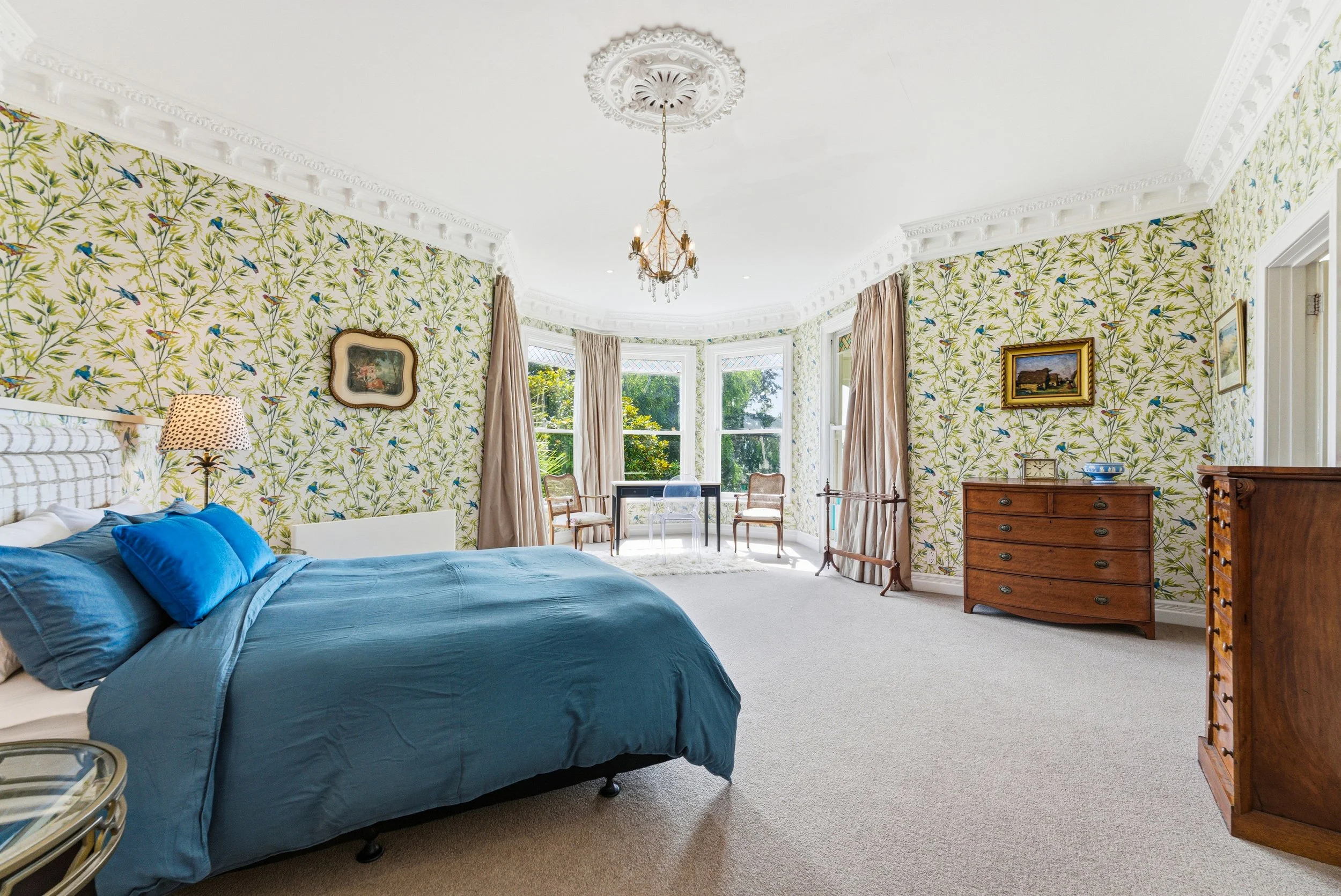 Bedroom with floral wallpaper, bay window with curtains, blue bedding, wooden furniture, chandelier, and artwork.