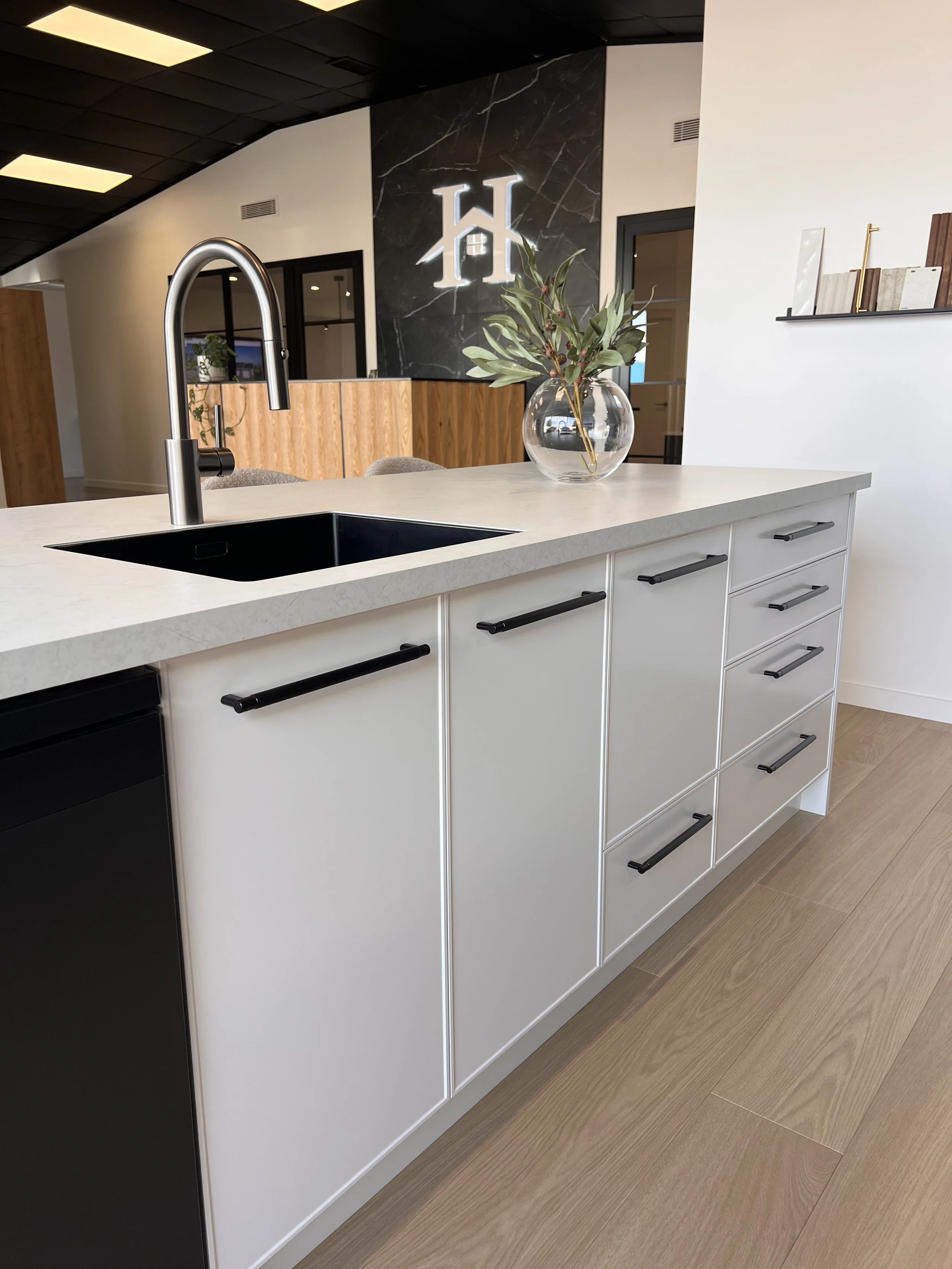 Modern kitchen island with white cabinets, black handles, a black sink, and a tall curved faucet. A glass vase with green leaves is on the counter. The background features a black marble wall with a white illuminated logo and a wooden panel.