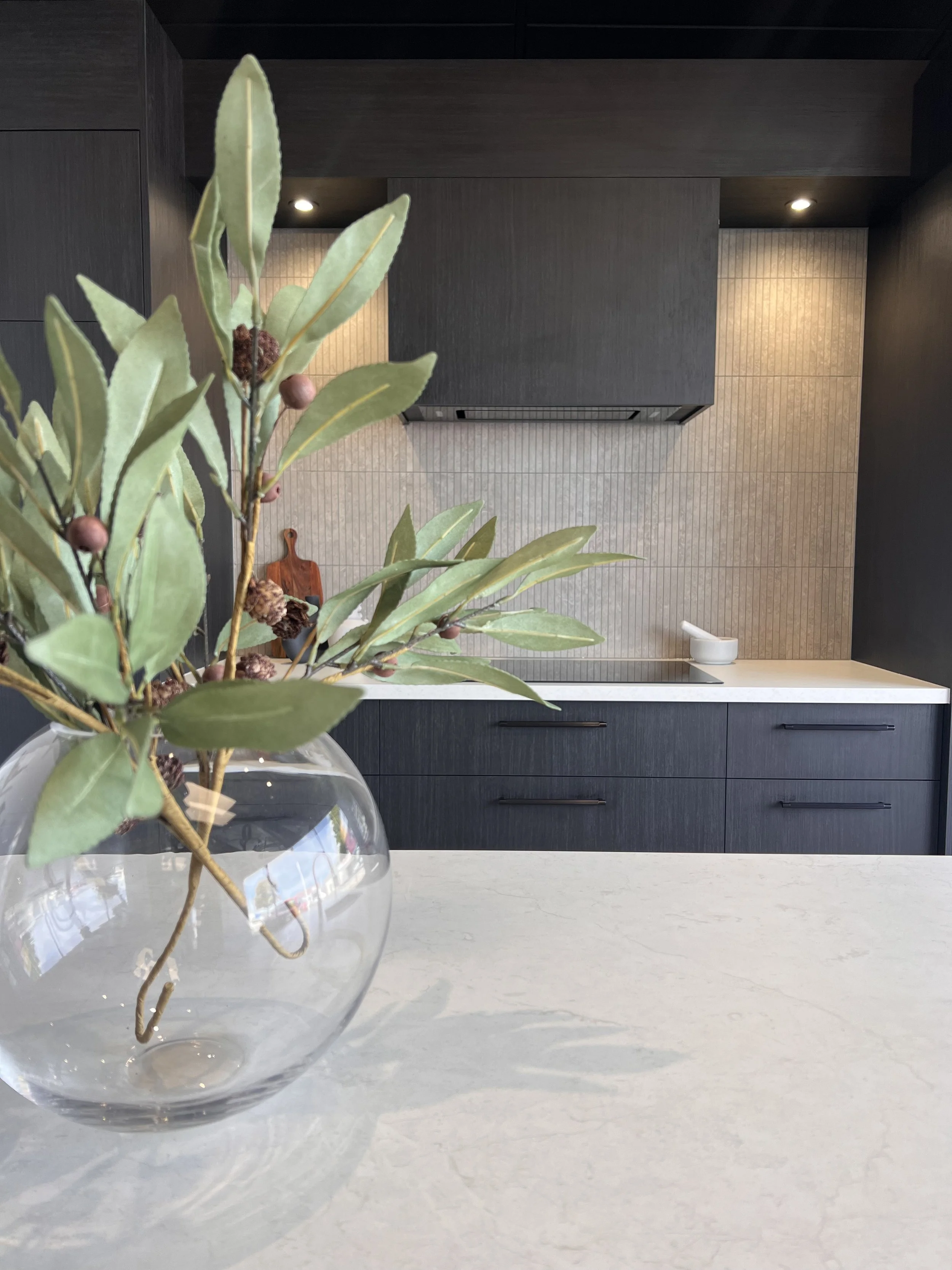 Close-up of a glass vase with green foliage and berries on a white countertop in a modern kitchen with dark cabinets and textured beige backsplash.