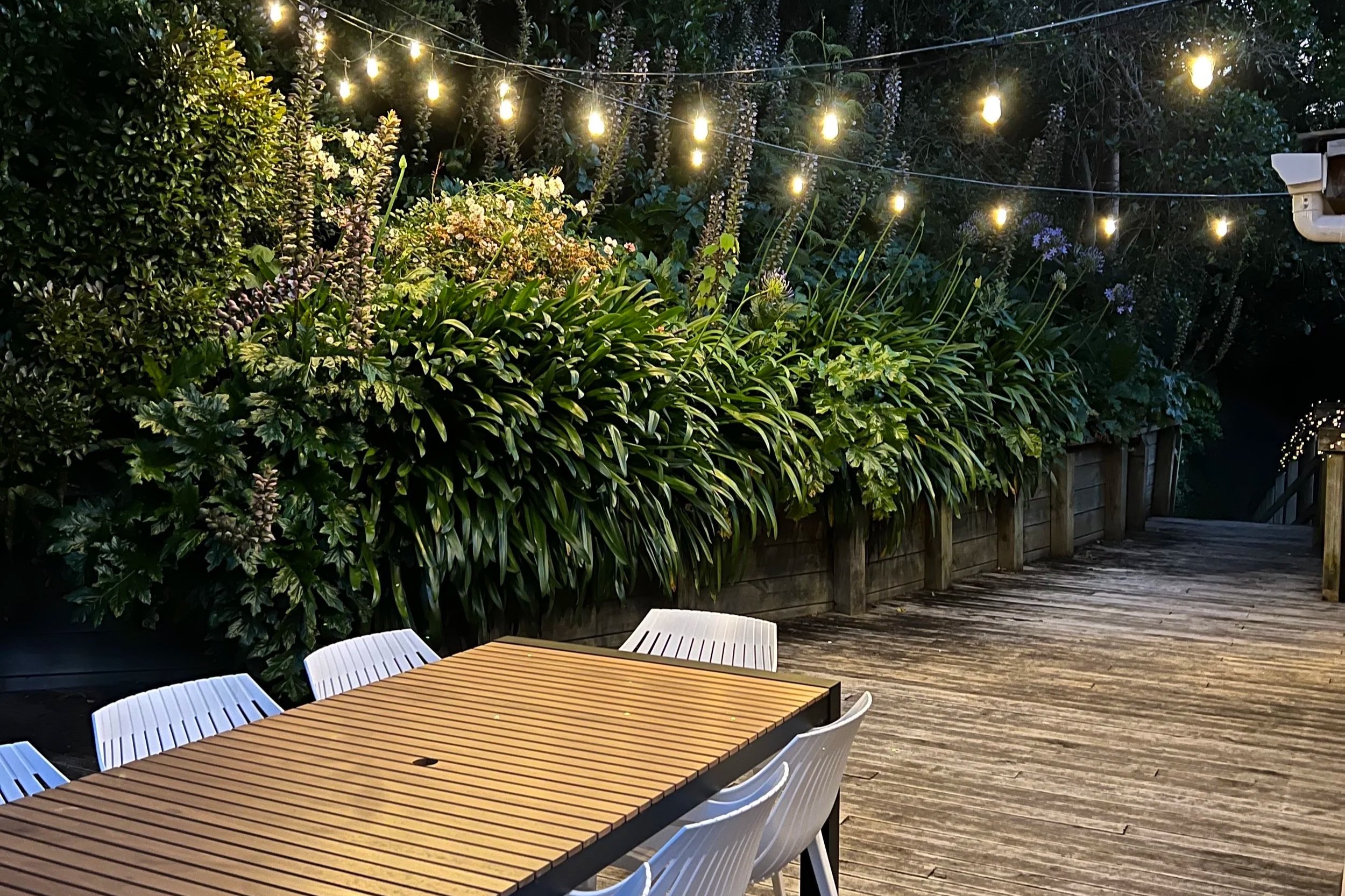 Outdoor patio scene at dusk with wooden table and white chairs, lush green bushes, and string lights hanging above.