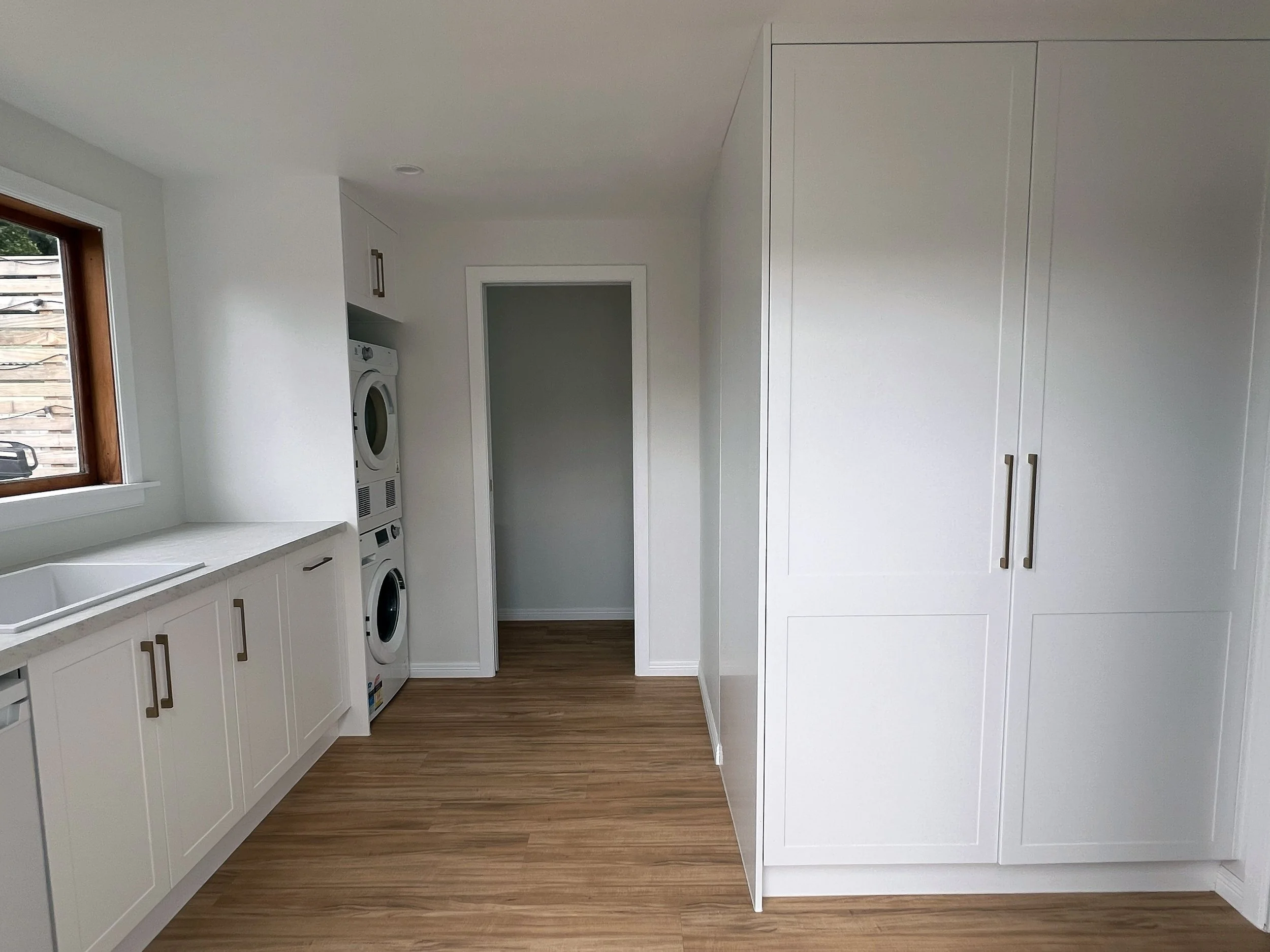 A laundry room with white cabinets, a window with wooden trim, a stacked washer and dryer, and wood flooring.