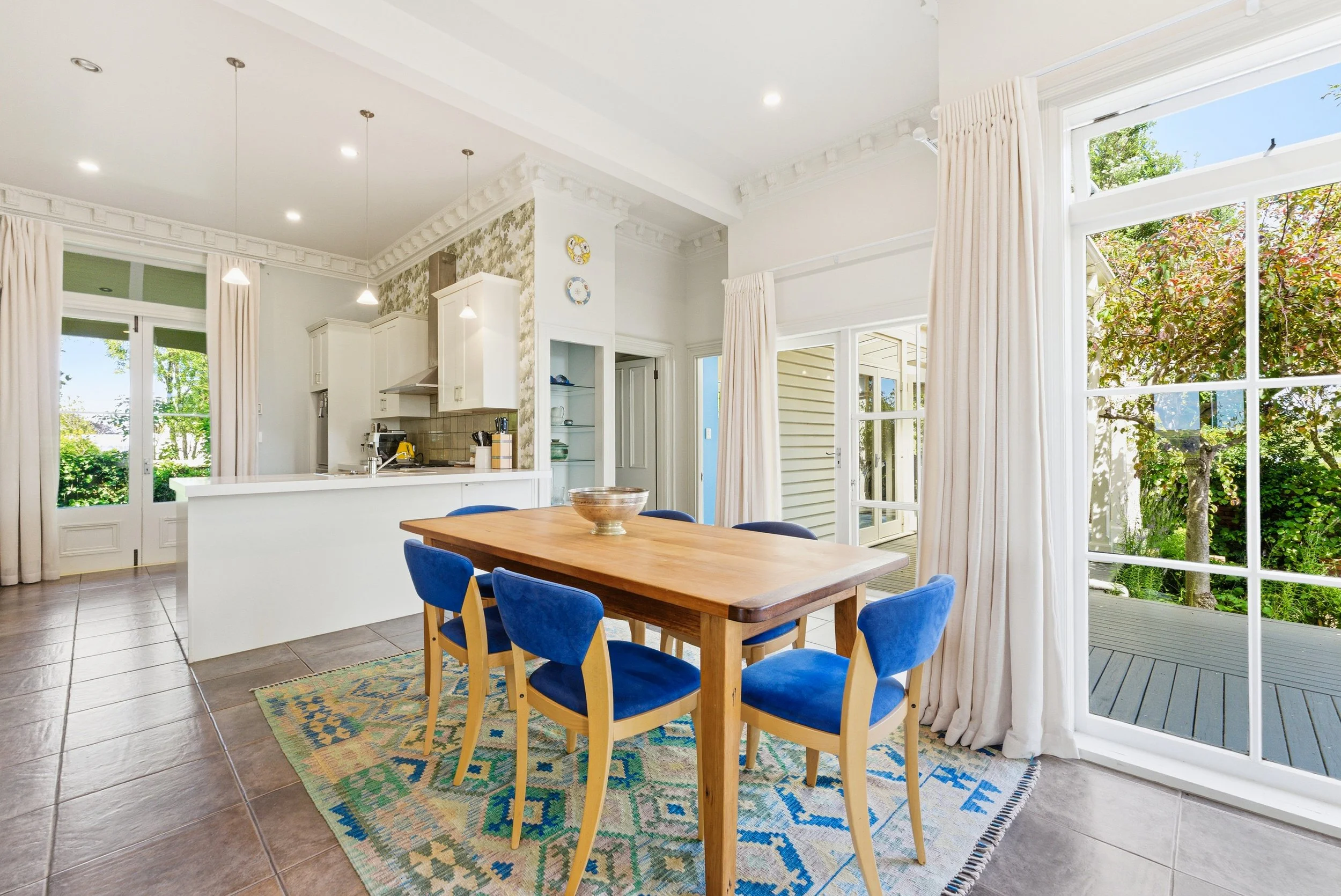 Bright dining area with a wooden table and blue upholstered chairs, adjacent to a kitchen with white cabinets and a breakfast bar, large windows with white curtains showing greenery outside.