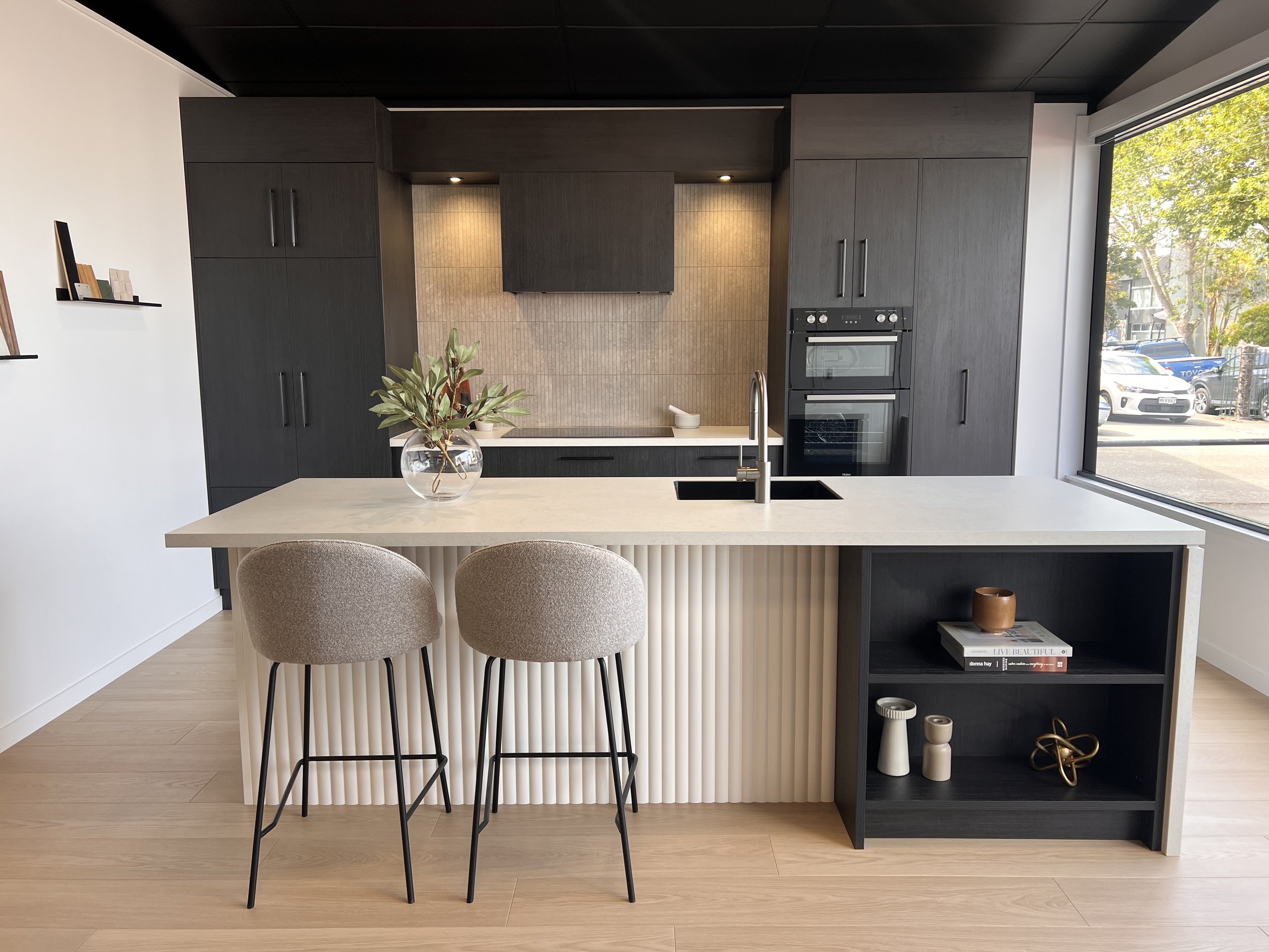 Modern kitchen with black cabinetry, a large white island with a built-in sink and beige barstools, and a large window overlooking a parking lot and trees.