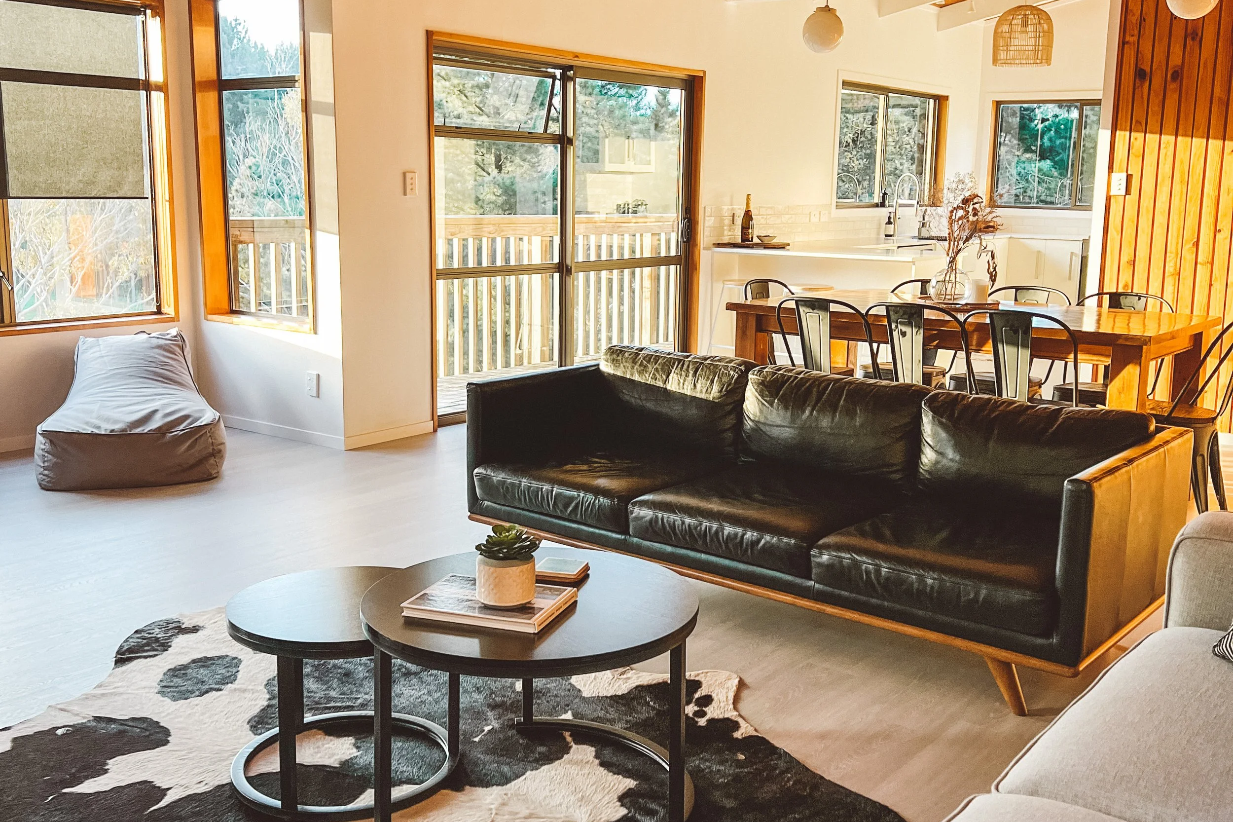 Living room with black leather sofa, two round coffee tables holding books and a small plant, beige armchair, cowhide rug, large windows, and a wooden dining table with chairs in an open kitchen area.