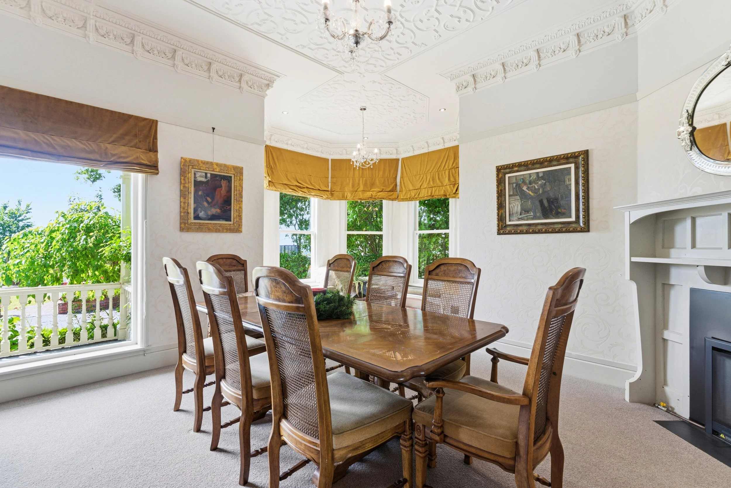 Elegant dining room with a large wooden table and ten wooden chairs. The room has ornate ceiling molding, two chandeliers, and a fireplace with a mirror above it. Large windows with gold curtains let in natural light, and there are framed paintings on the walls.