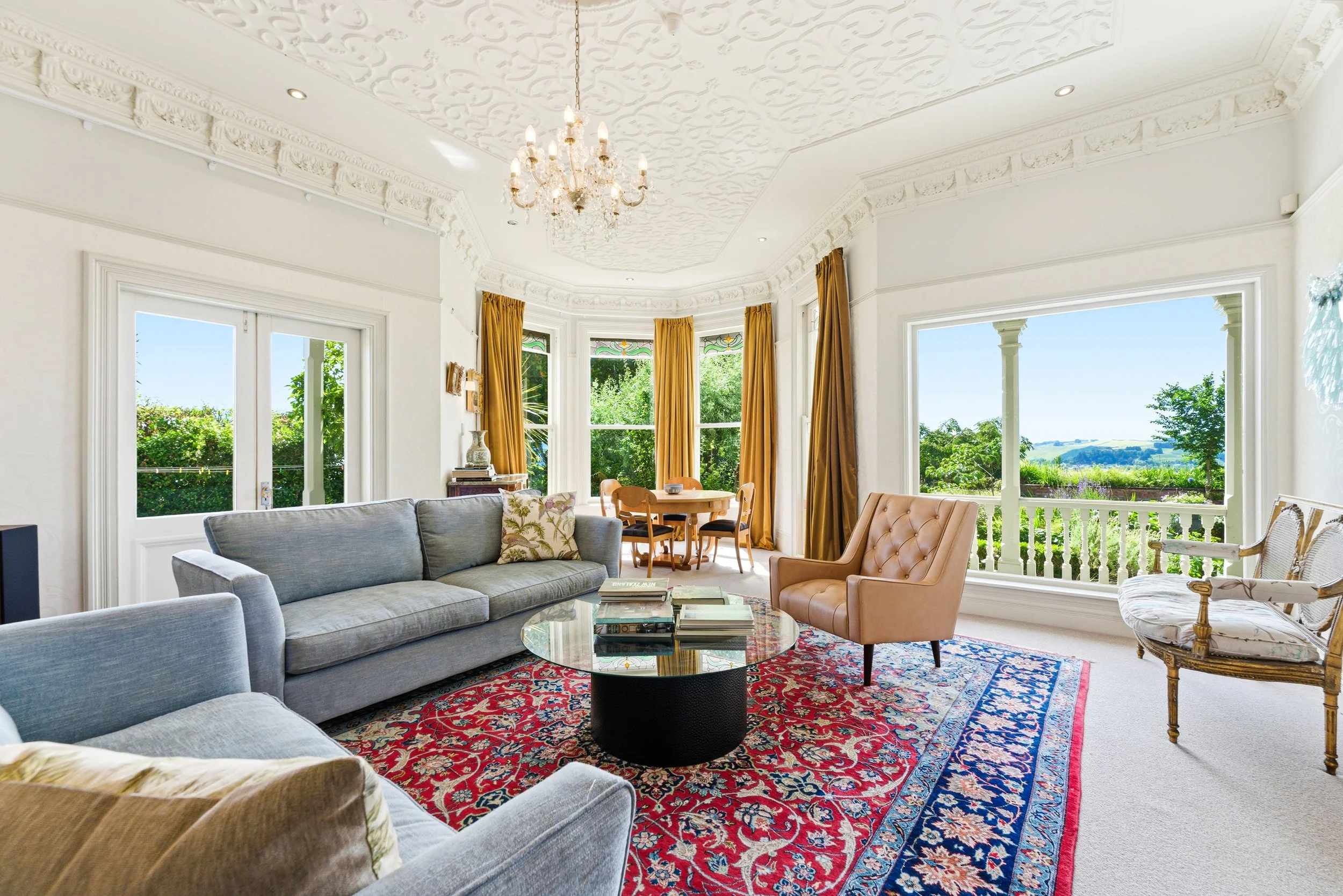 Living room with large windows, ornate white ceiling, chandelier, gray sofa, pink armchair, patterned rug, round coffee table with books, gold curtains, and scenic outdoor view