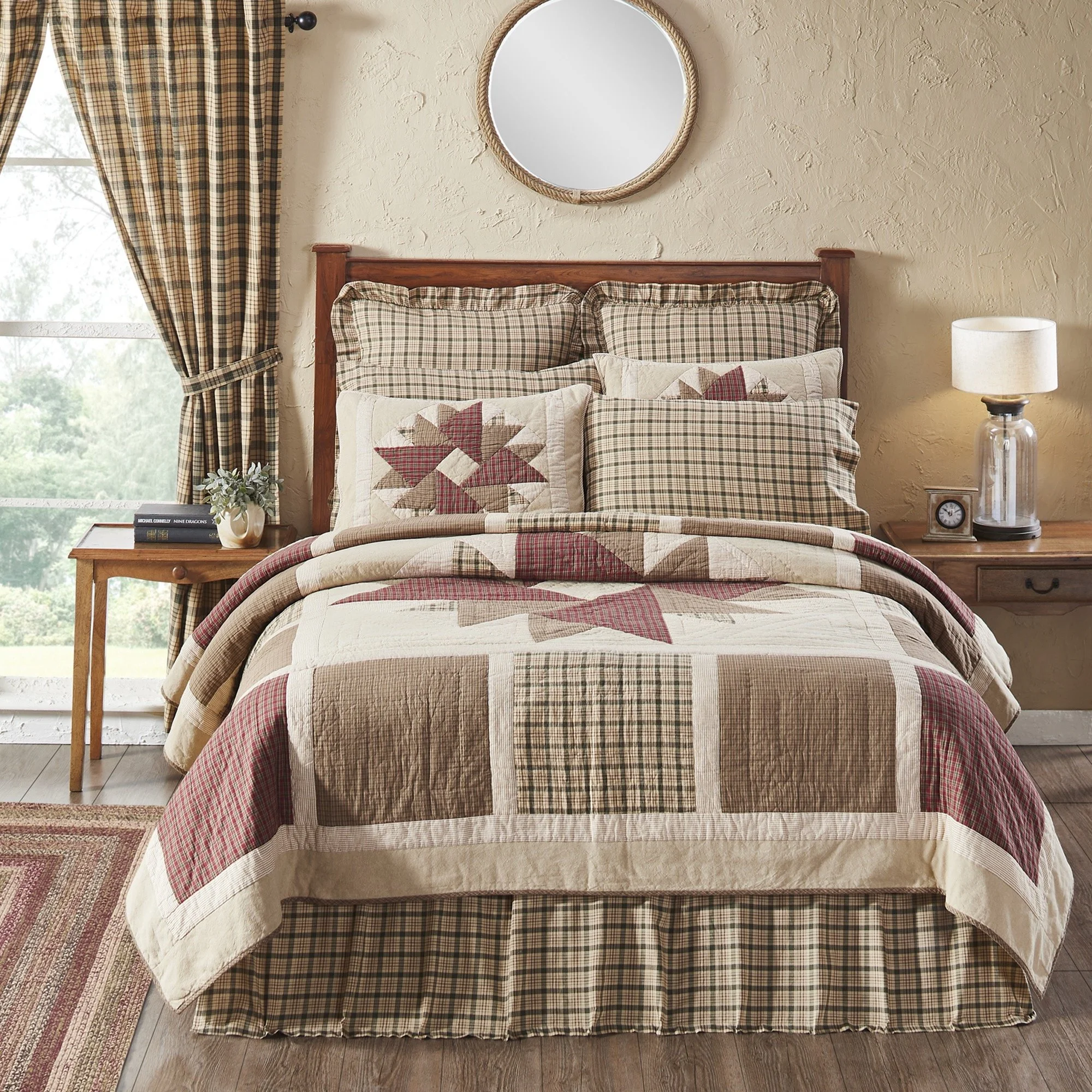 A bed with a patchwork quilt and plaid pillows, situated beside a window with plaid curtains, in a cozy bedroom with beige walls, a round mirror, a table with a lamp, clock, and plants.