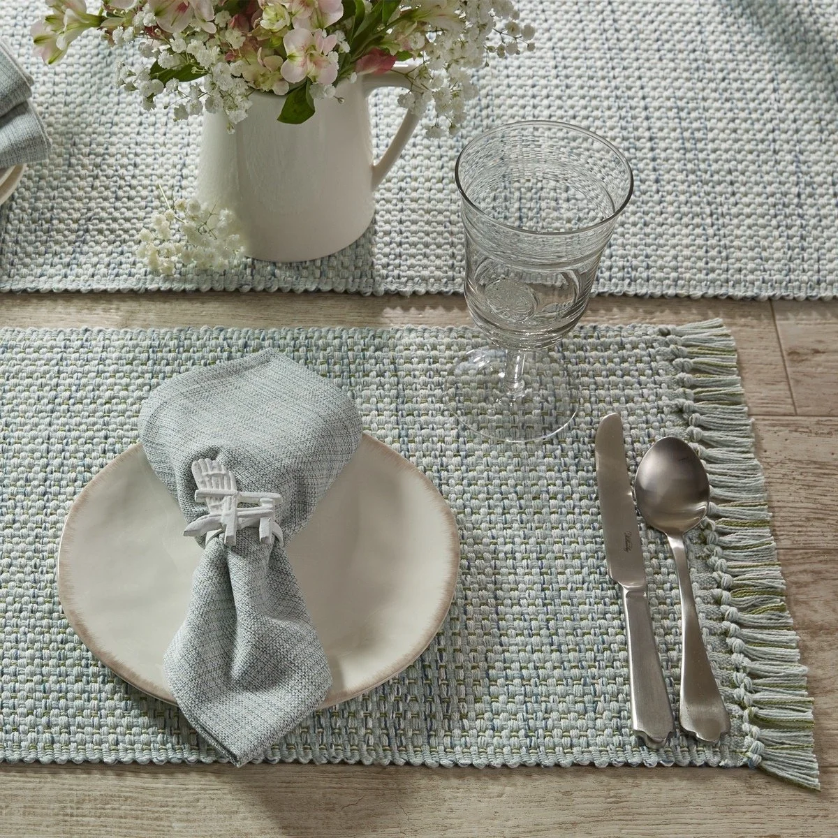 Table setting with a white ceramic plate, a folded gray cloth napkin secured with a white clip, a knife and spoon on a textured woven placemat, a water glass, and a white vase with pink and white flowers on a light wooden table.