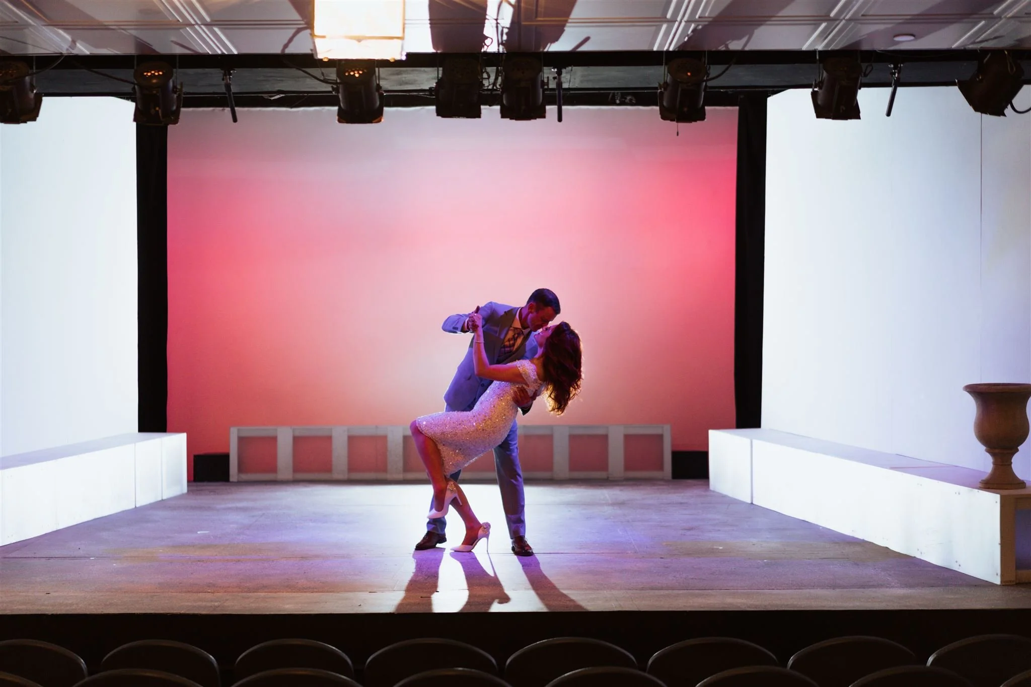 A man and woman dance on a stage with a pink and orange gradient backdrop. The woman is wearing a shimmering dress and heels, while the man is in a suit. They are dancing closely, with the man dipping the woman.