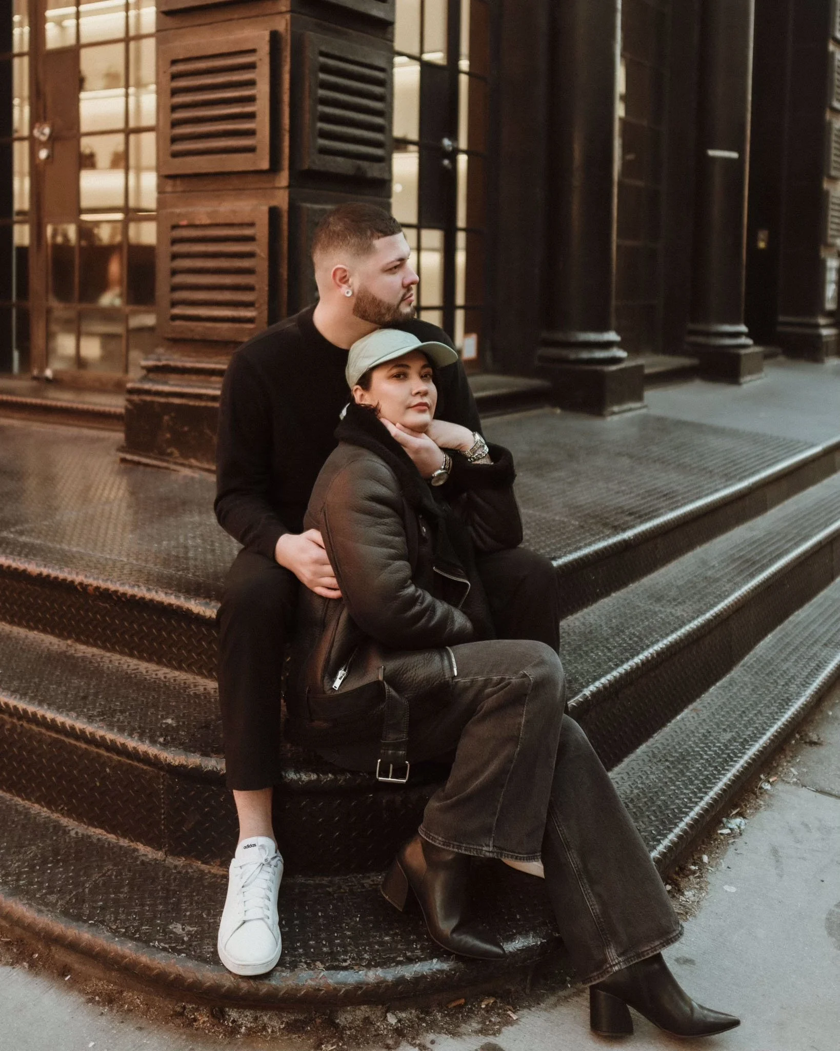 A man and woman sitting on metal stairs outside a building with wooden shutters and glass windows, with the man behind and the woman in front. The woman wears a baseball cap, leather jacket, gray jeans, and black boots. The man wears a black long-sleeve shirt, black pants, and has short hair and a beard.