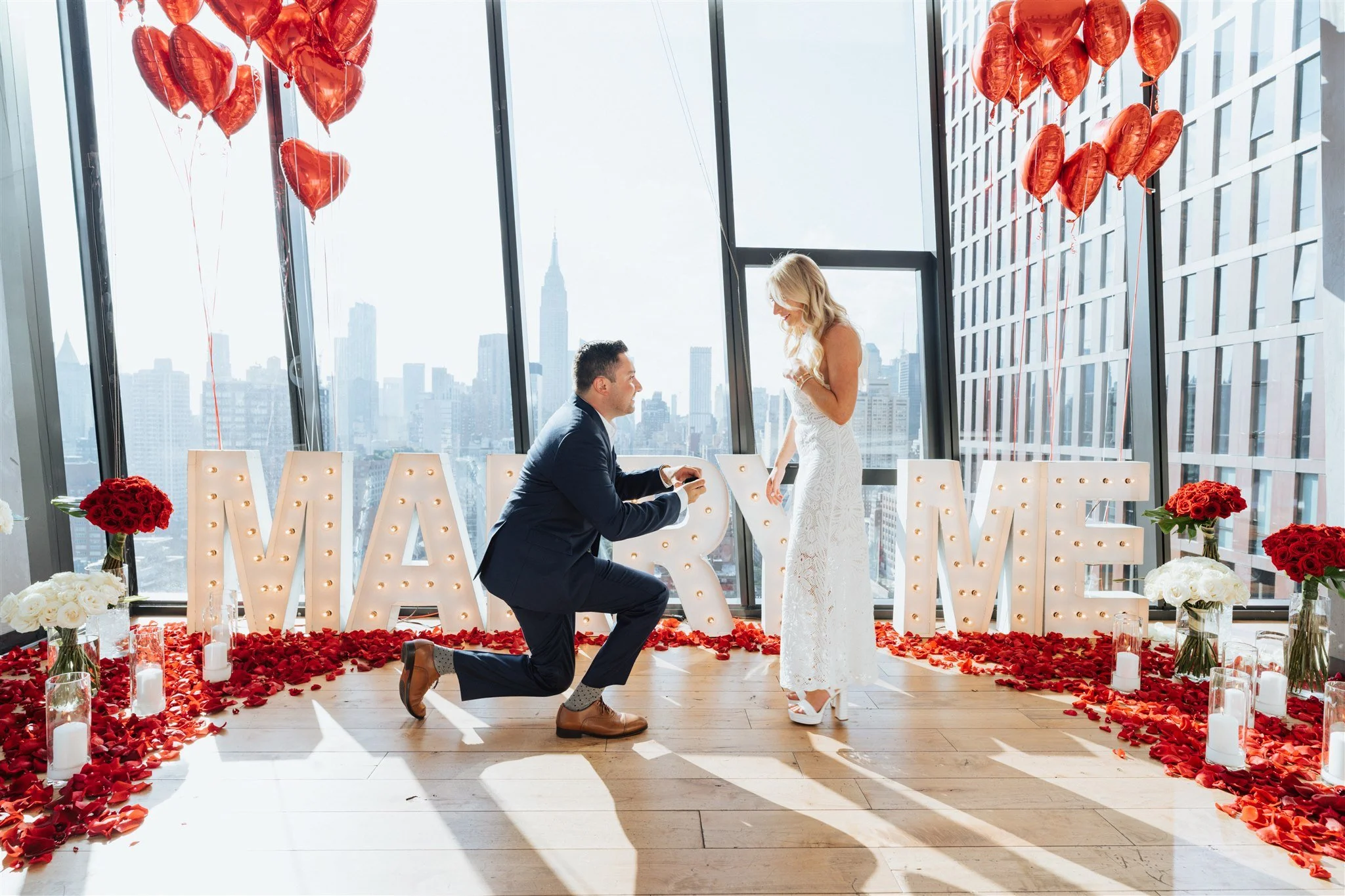 Man proposing on NYC rooftop with 'Marry Me' marquee and red rose petals