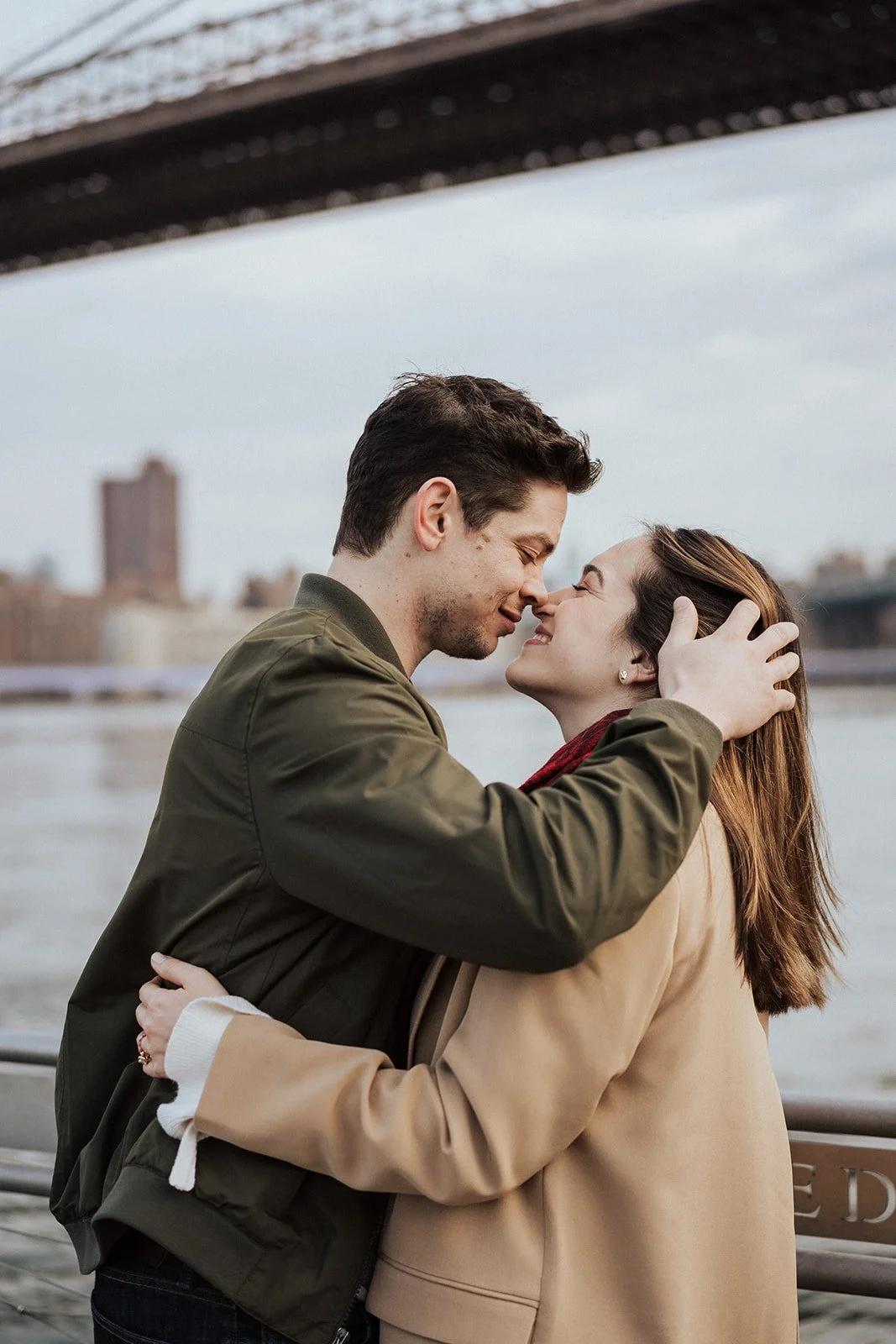 A romantic moment between a man and woman embracing near a river, under a bridge, with city buildings in the background.