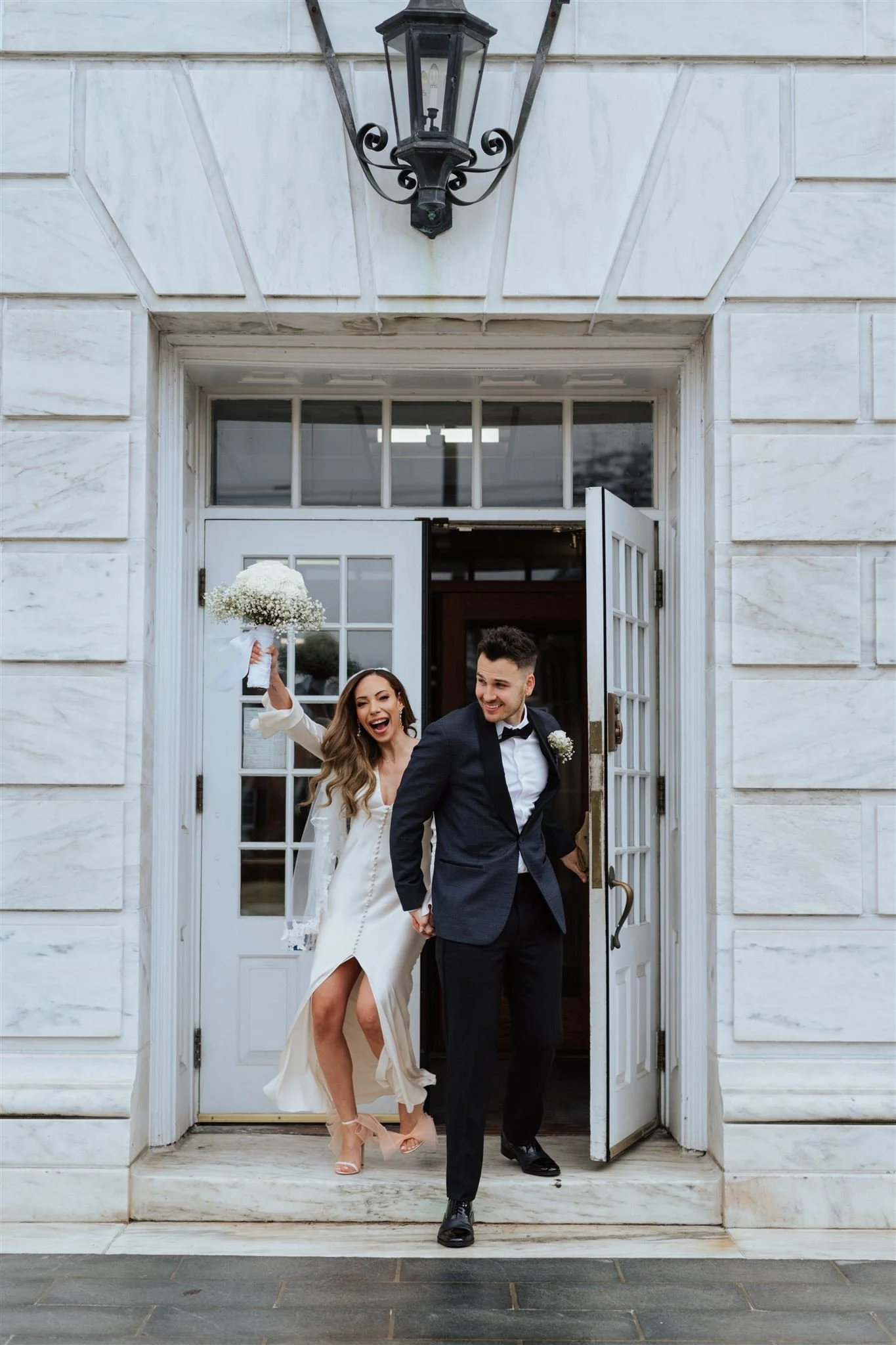 A joyful bride and groom exiting a building through double glass doors, holding hands. The bride, wearing a white dress, is happily throwing a bouquet, and the groom, in a tuxedo, smiles at her.