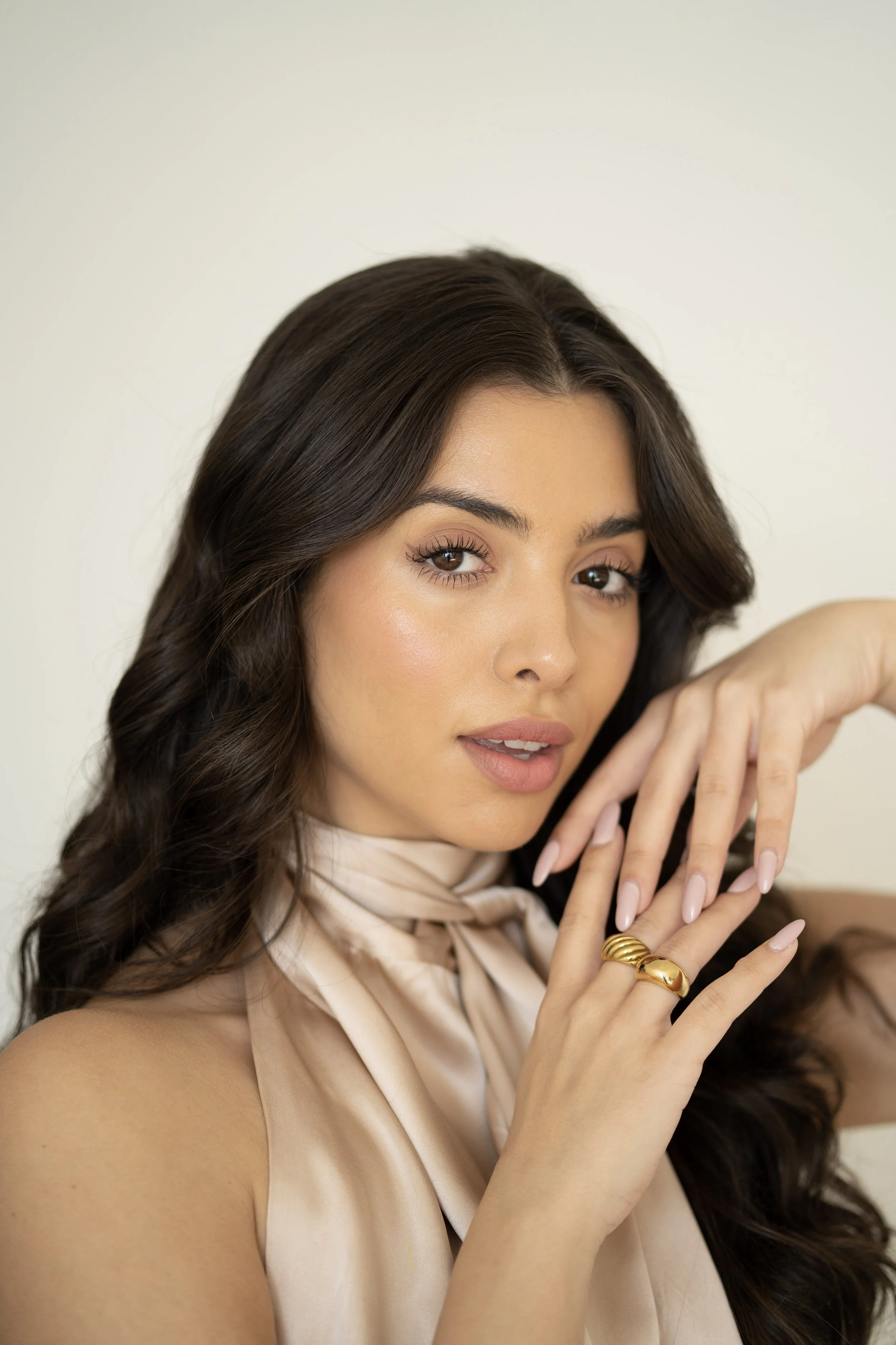 Close-up portrait of a young woman with long, wavy brown hair, wearing a silky beige blouse, gold rings, and showing well-manicured nails, posing against a plain white background.