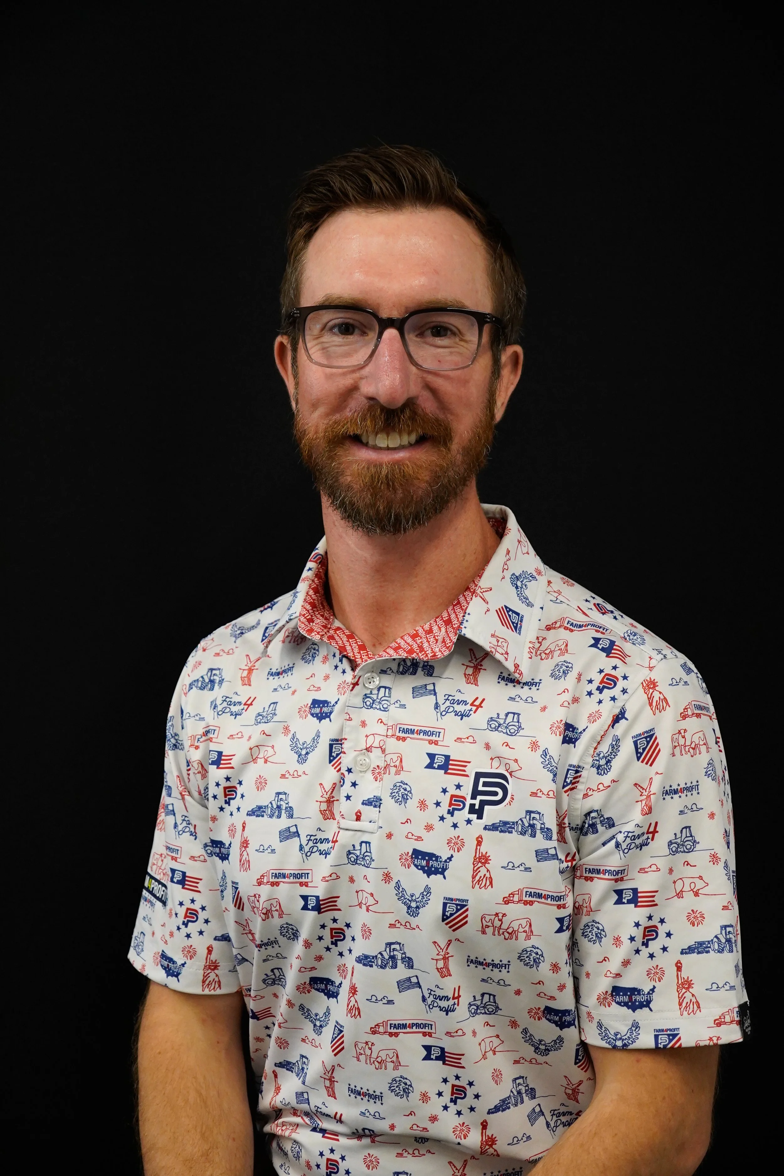 A smiling man with glasses, a beard, and short brown hair, wearing a red, white, and blue patriotic themed shirt with farm and American motifs, against a black background.