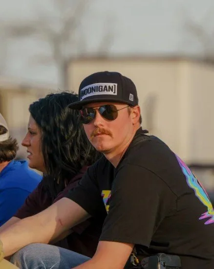 A man with sunglasses, a mustache, wearing a black cap and t-shirt, sitting outdoors with a woman with dark hair, and others partly visible.