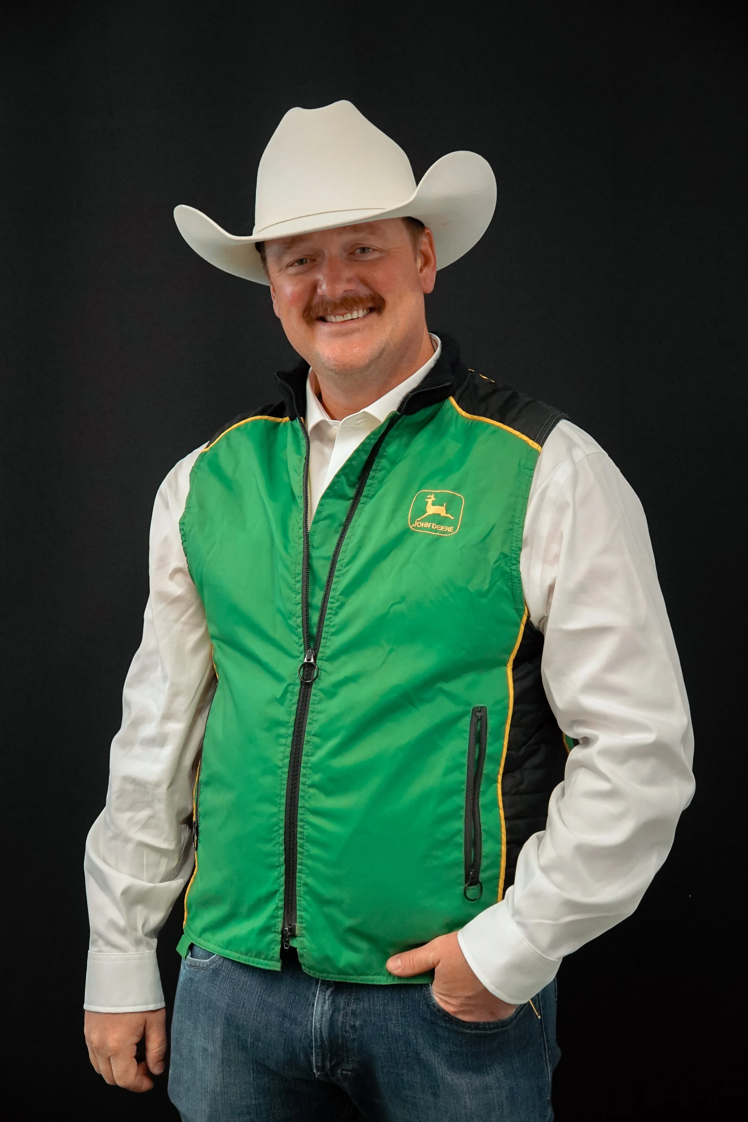 Man wearing a white cowboy hat, green and black John Deere vest, white shirt, and jeans, standing against a black background smiling at the camera.