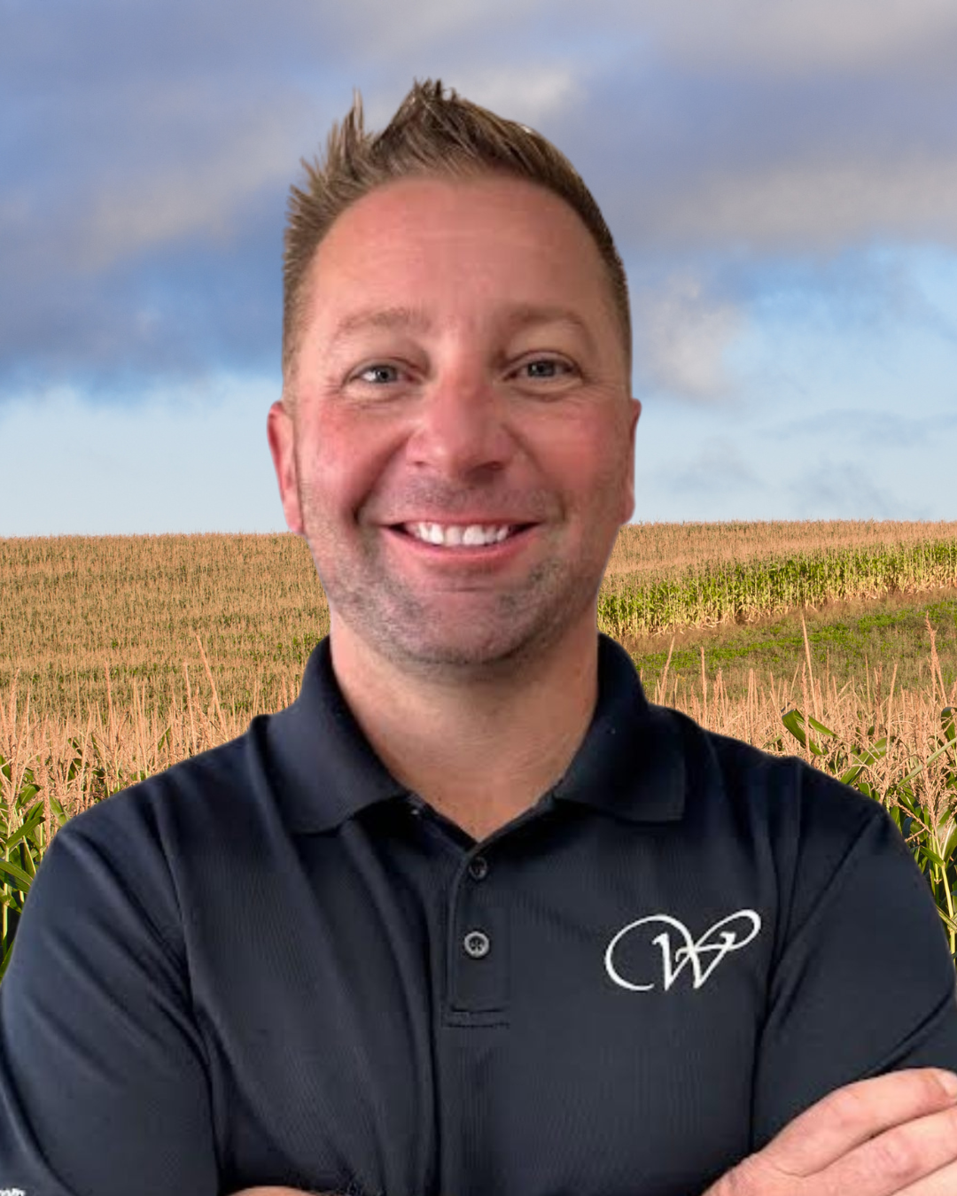 A smiling man with short brown hair wearing a black polo shirt with a white logo, standing in front of a field of crops under a partly cloudy sky.
