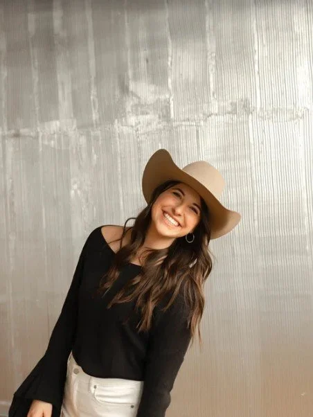 A young woman smiling and wearing a large beige floppy hat and black top, standing in front of a light gray textured wall.